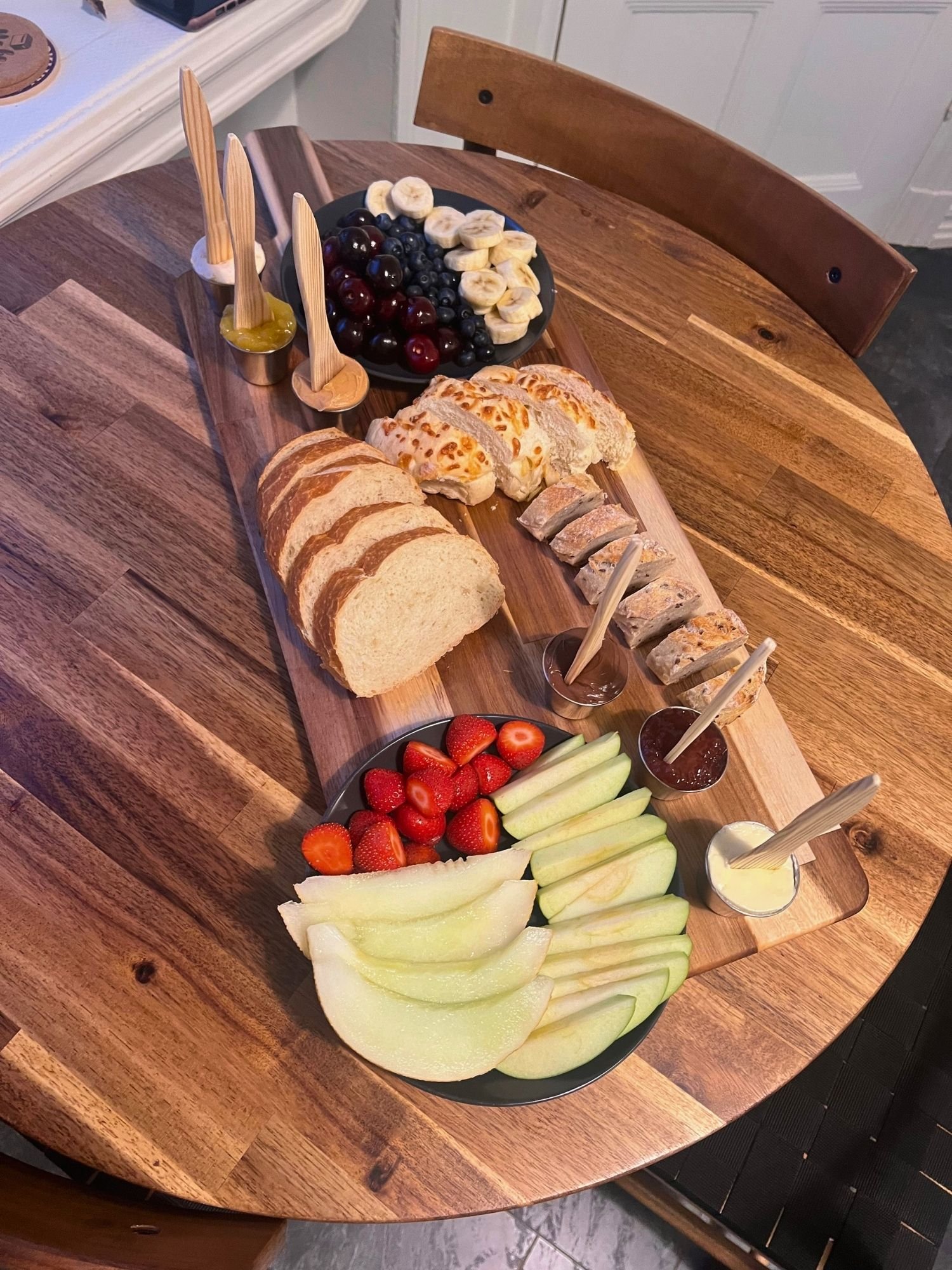 High-angle shot of a long wooden serving board on a round wooden table set with sliced bread and fresh fruit.