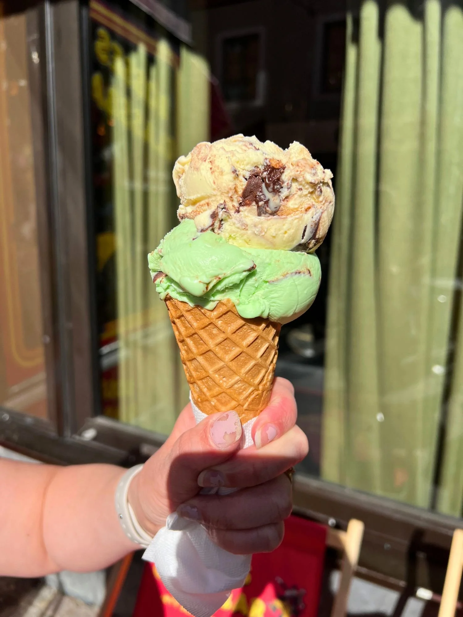 A waffle cone with a scoop of mint chocolate and a scoop of cookie dough ice cream held up in front of a shop window.