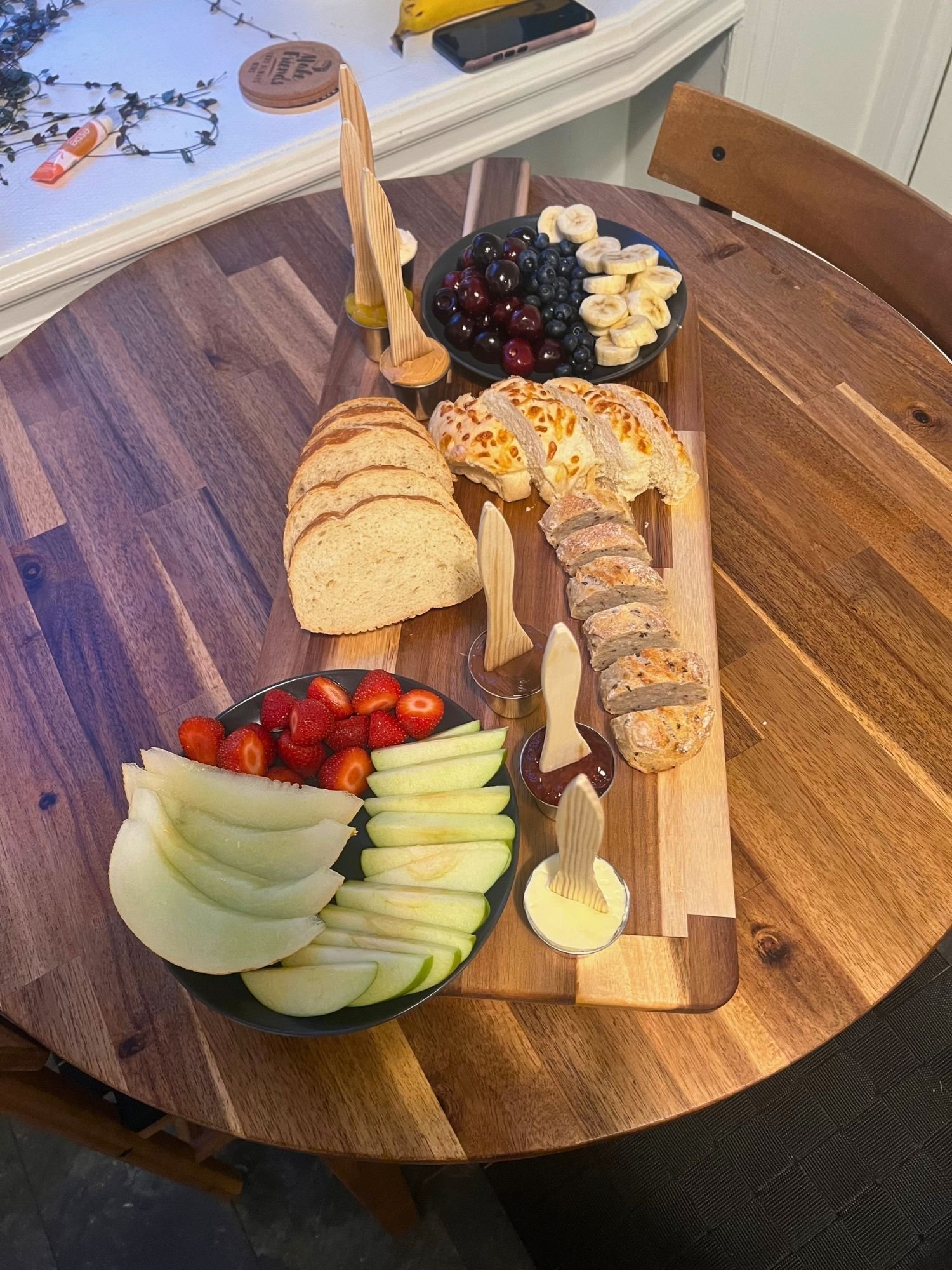 A rectangular wooden charcuterie board rests vertically across a round wooden table, filled with fruit, sliced bread, and spreads.