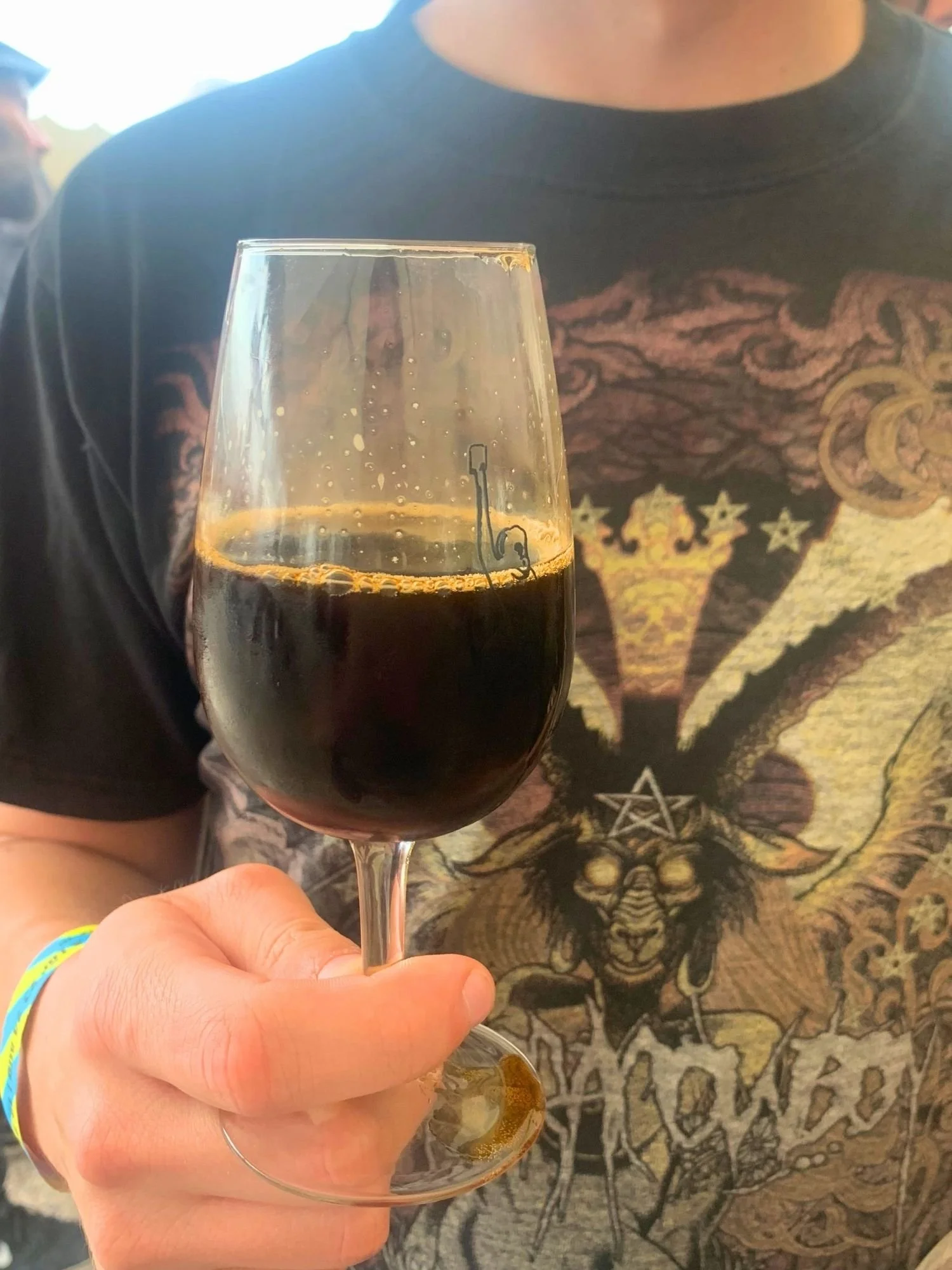 A hand holds up a glass of dark stout beer with a minimal head. The person holding the glass wears a black graphic t-shirt featuring an ornate, dark illustration, along with a yellow and blue festival wristband.