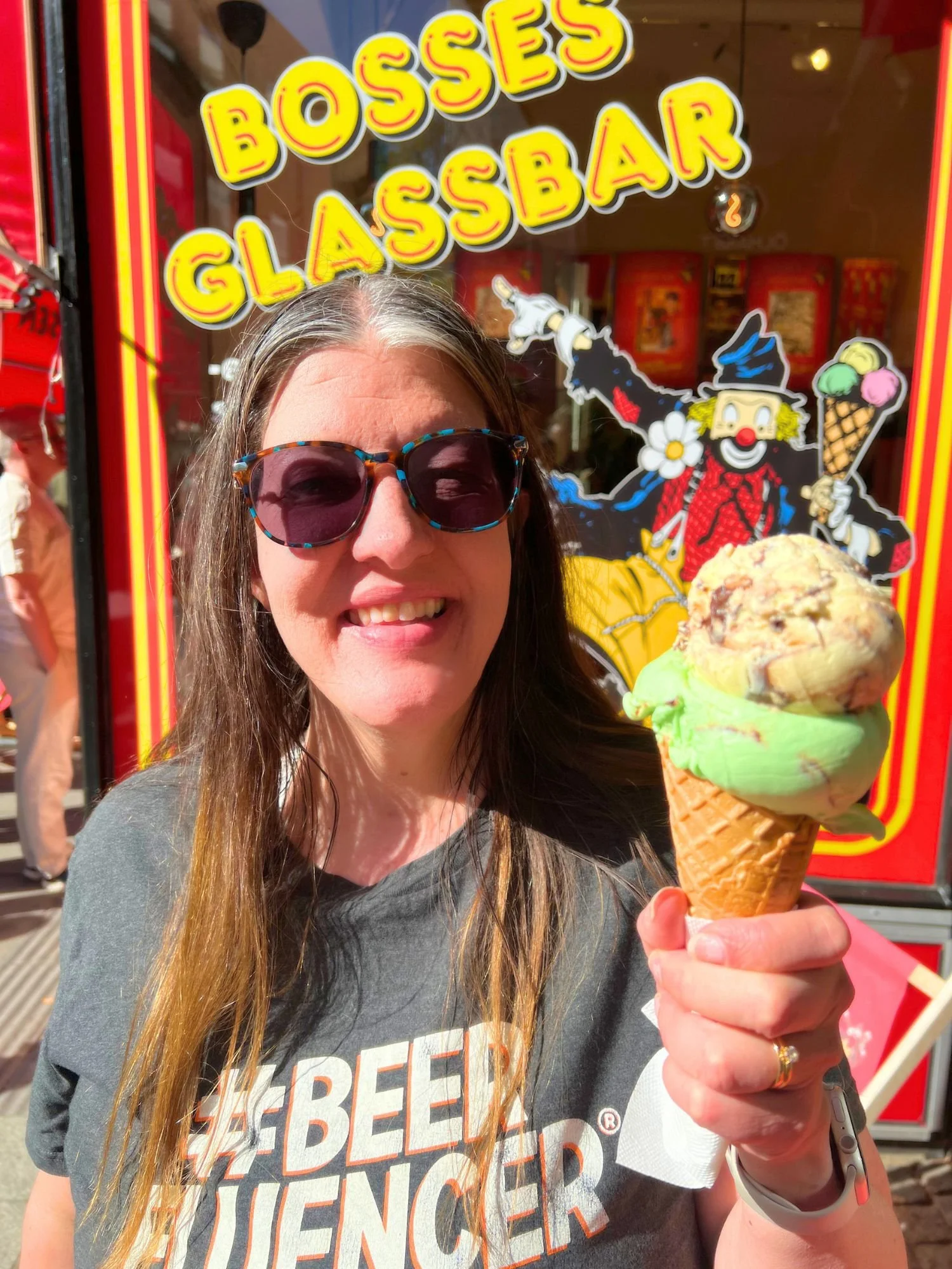 A smiling woman with long brown hair, grey-streaked roots, and dark tortoiseshell sunglasses holding a double-scoop ice cream cone.
