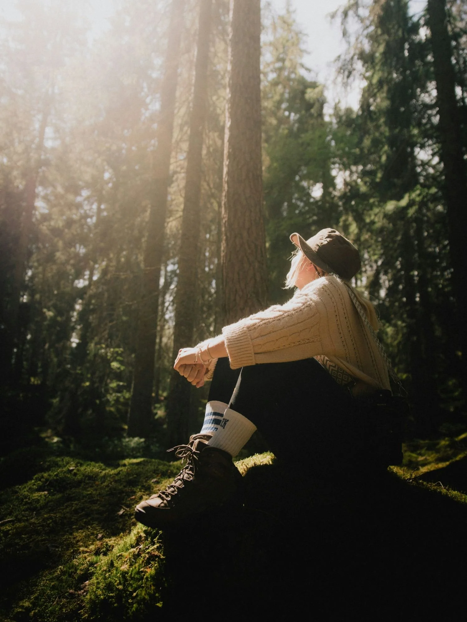 A woman wearing a brown bucket hat, a thick beige cable-knit sweater, dark pants, white socks with blue stripes, and sturdy hiking boots sits resting in profile on a large, moss-covered rock in Tiveden National Park.