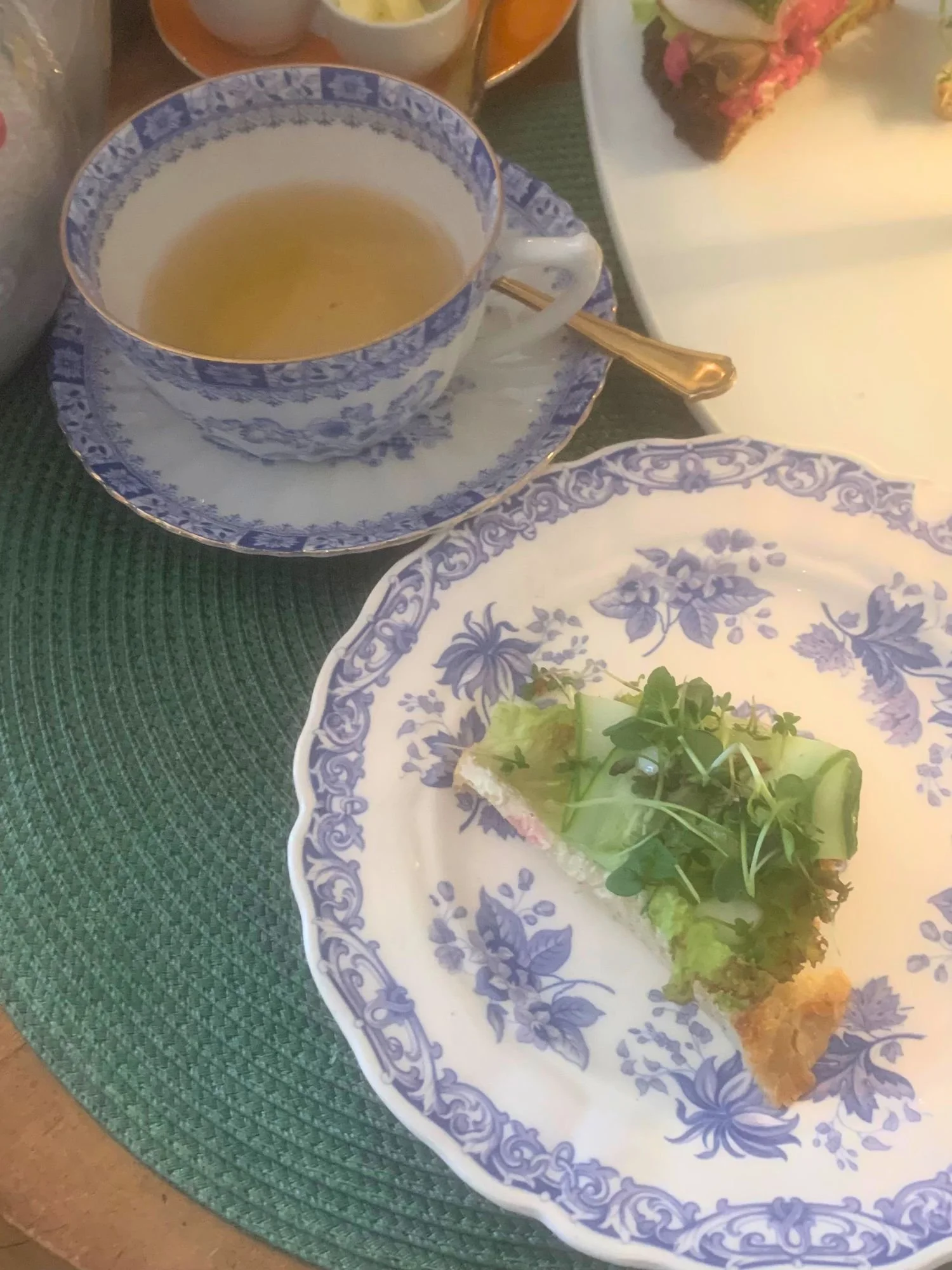 Tea service at Alice och Kattens Tesalong in Lund. A triangular slice of bread topped with a light spread, fresh lettuce, thinly sliced cucumber ribbons, and delicate microgreens, served on a white plate with an intricate blue floral pattern.