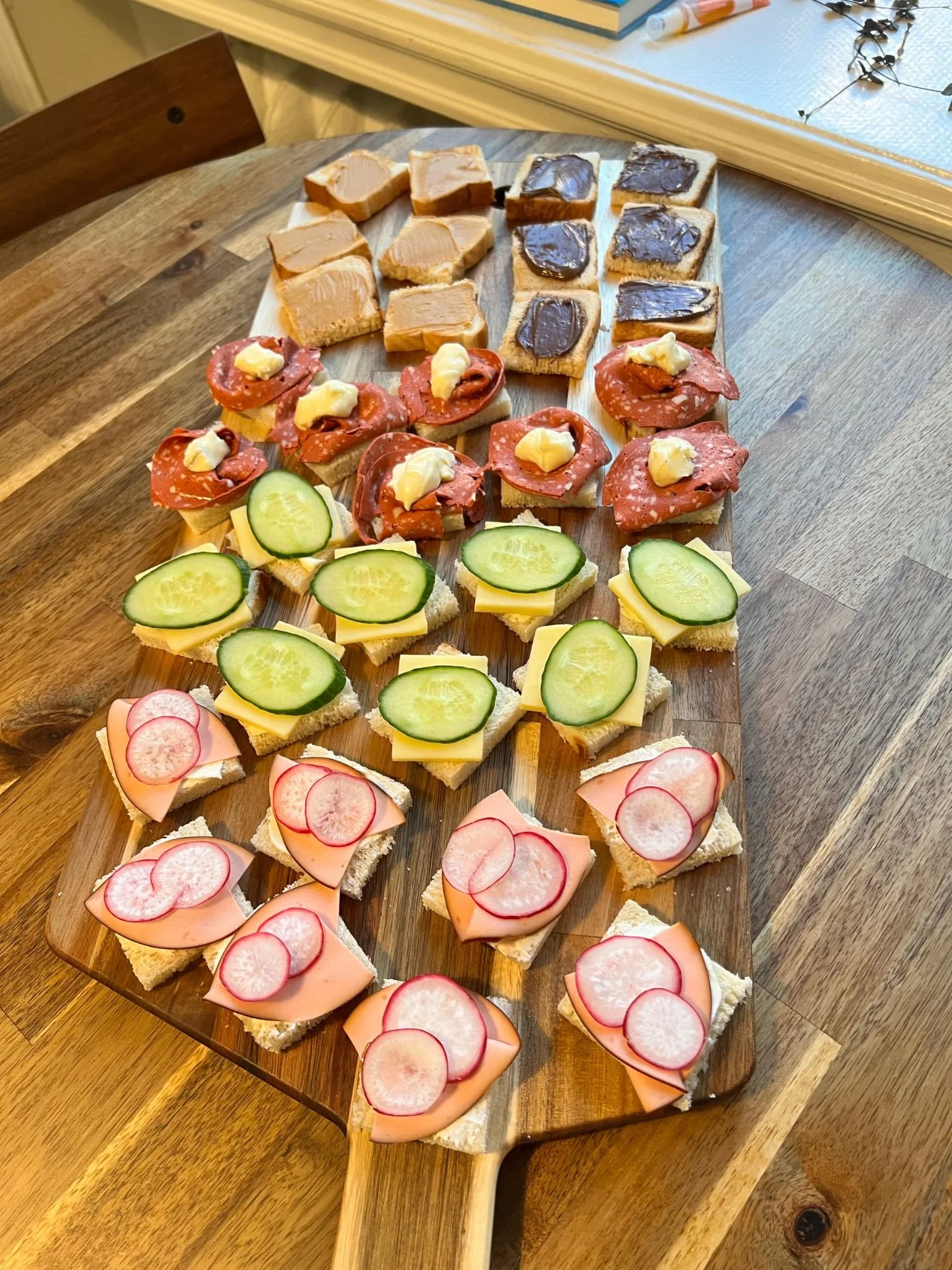 A wooden serving board on a round wooden table filled with rows of mini square open-faced sandwiches with sweet and savory toppings.