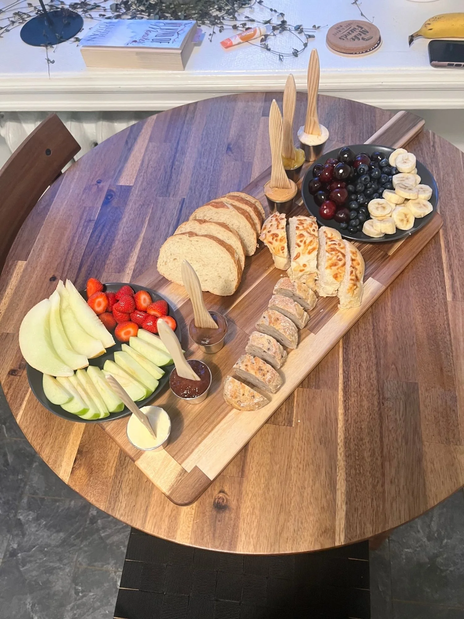 A rectangular wooden serving board lies diagonally across a round wooden table, laden with sliced bread, fruit plates, and small cups of spreads.