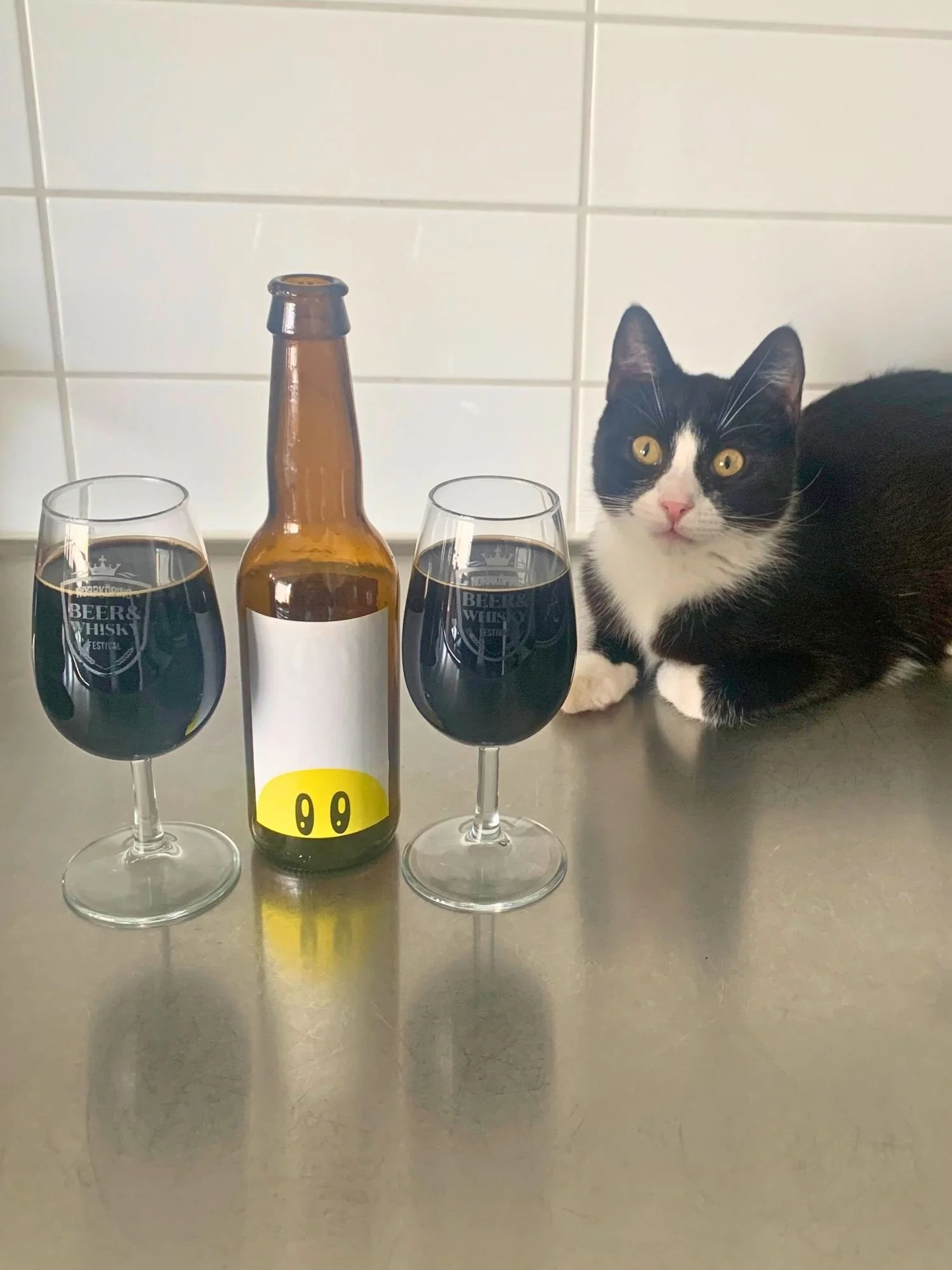A brown beer bottle with a minimalist label featuring half of a yellow smiley face between two glasses filled with dark imperial stout on a stainless steel surface. The glasses are printed with the text "Norrköping Beer & Whisky Festival".