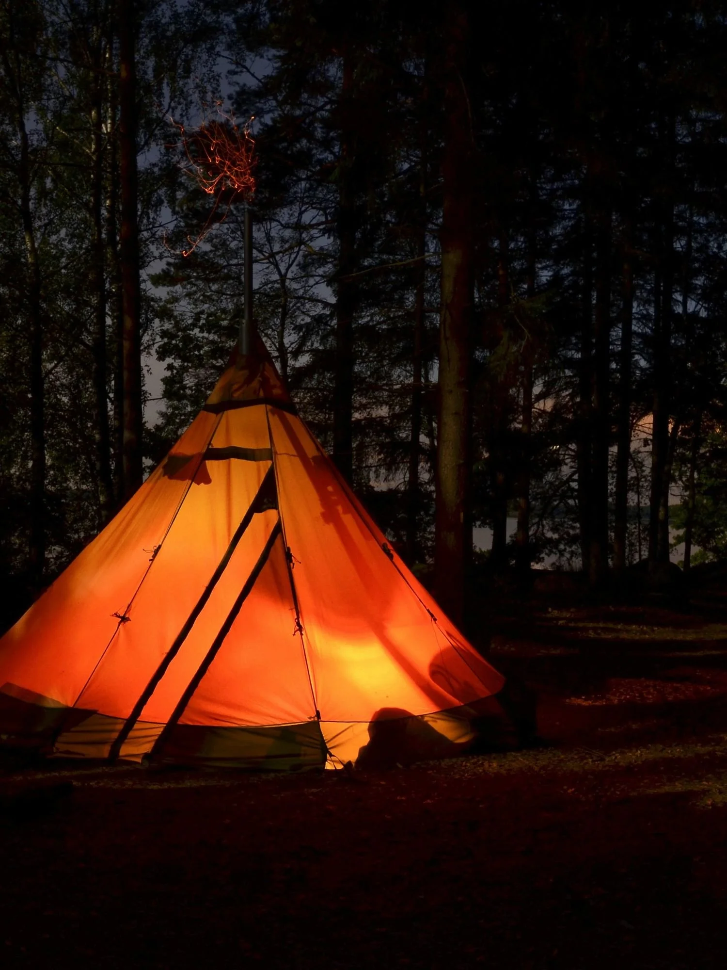 A large, conical tipis-style tent glows with a vibrant orange light from within during a dark night in a forest. The tent features a prominent stove pipe extending from the top peak, with glowing orange sparks faintly trailing into the air above it.