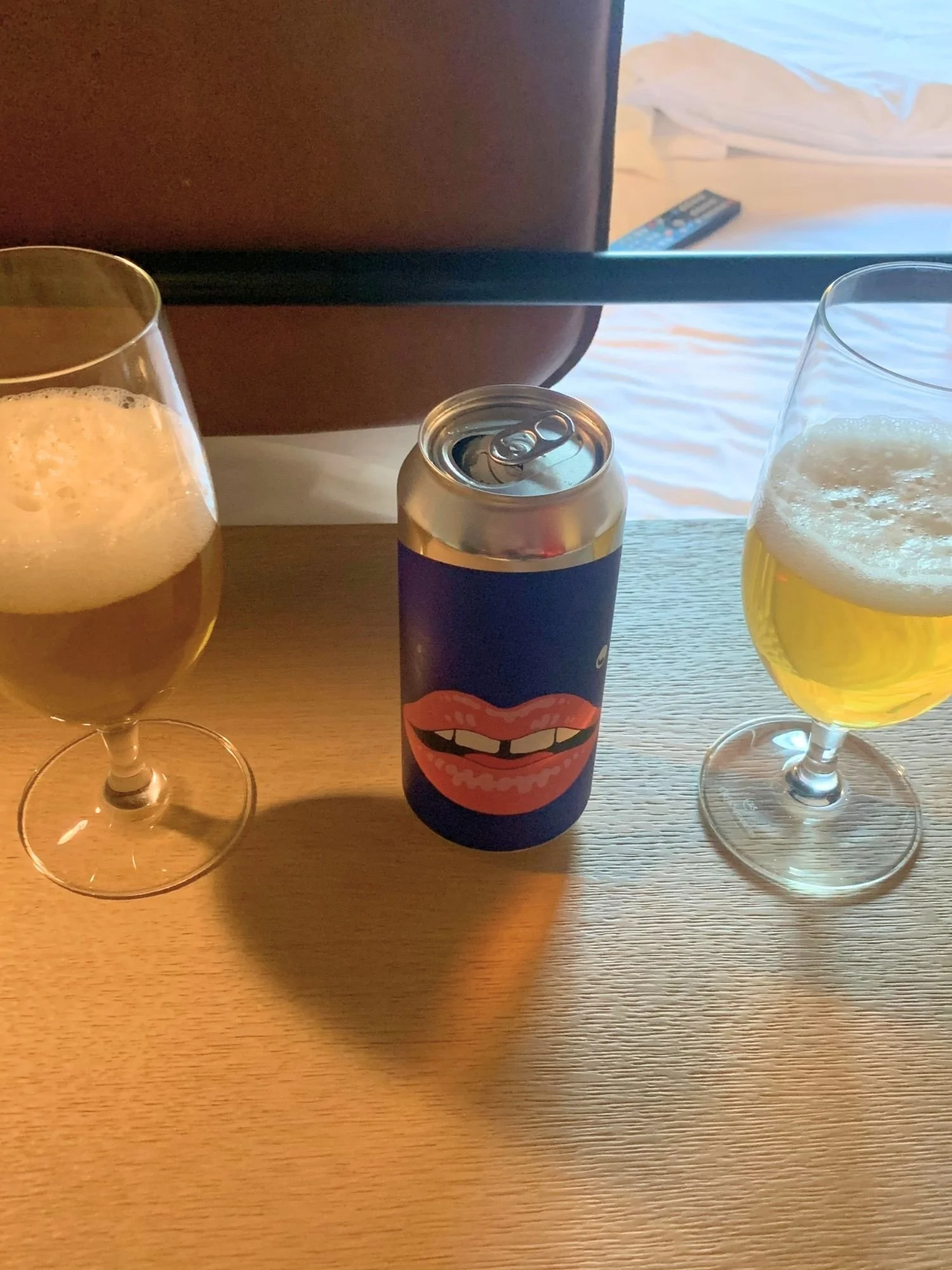 An open dark blue beer can featuring a graphic of smiling pink lips with visible teeth sits on a wooden desk, flanked by two stemmed glasses filled with golden beer topped with white foam.