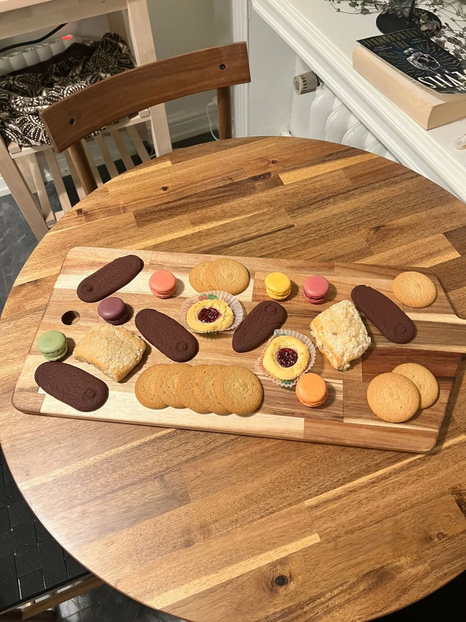A wooden serving board on a round wooden table, filled with an assortment of cookies.
