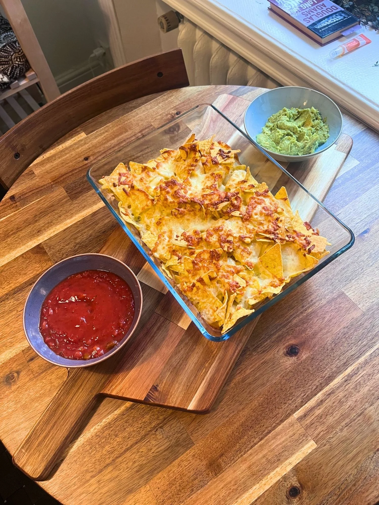 A large glass baking dish filled with melted cheese-covered tortilla chips, served on a wooden table alongside bowls of red salsa and guacamole.