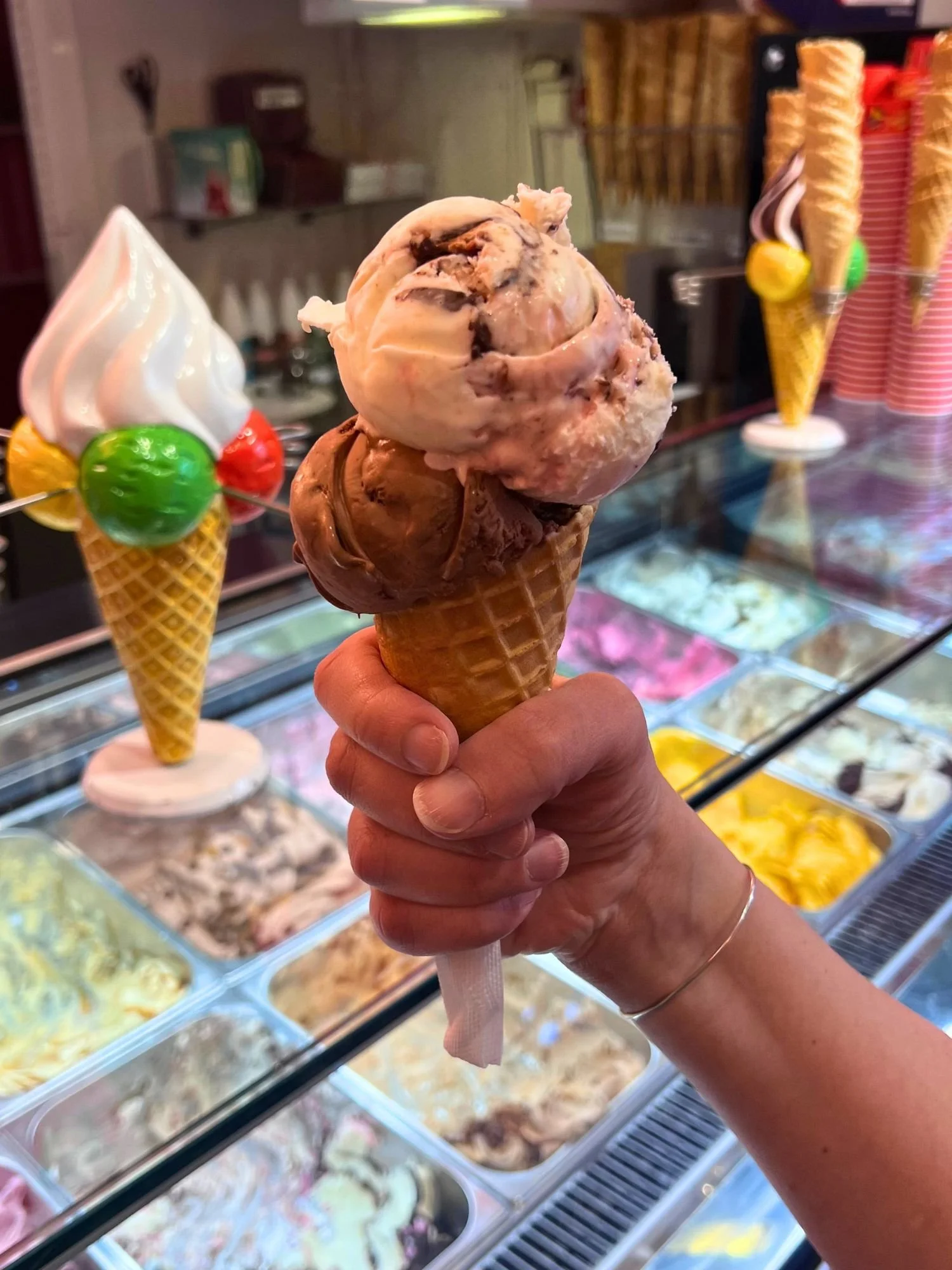 A hand holding a waffle cone with two scoops of ice cream, chocolate and cherry, inside an ice cream shop.
