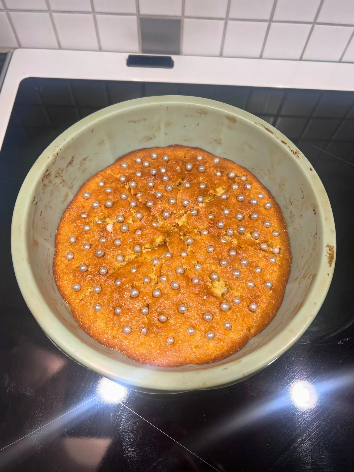 Golden orange cake topped with silver sugar pearls, baked in a round silicone cake pan on a black stovetop.