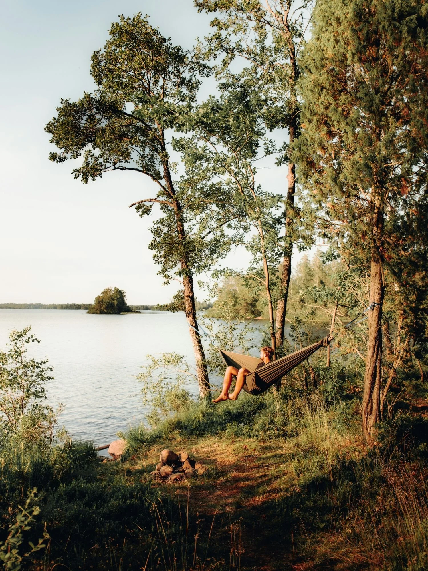 A person relaxes in a dark green camping hammock strung between two trees on a grassy, sunlit shoreline overlooking a wide, calm lake.
