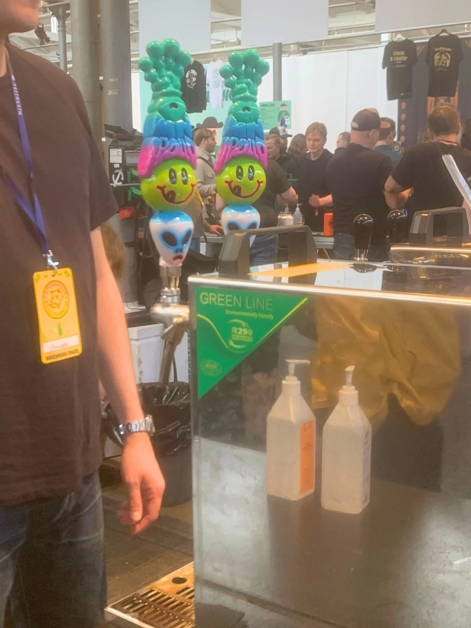 Two colorful, custom beer tap handles featuring stacked alien heads, yellow smiley faces, pink branding, and green textured tops stand on a stainless steel bar counter.