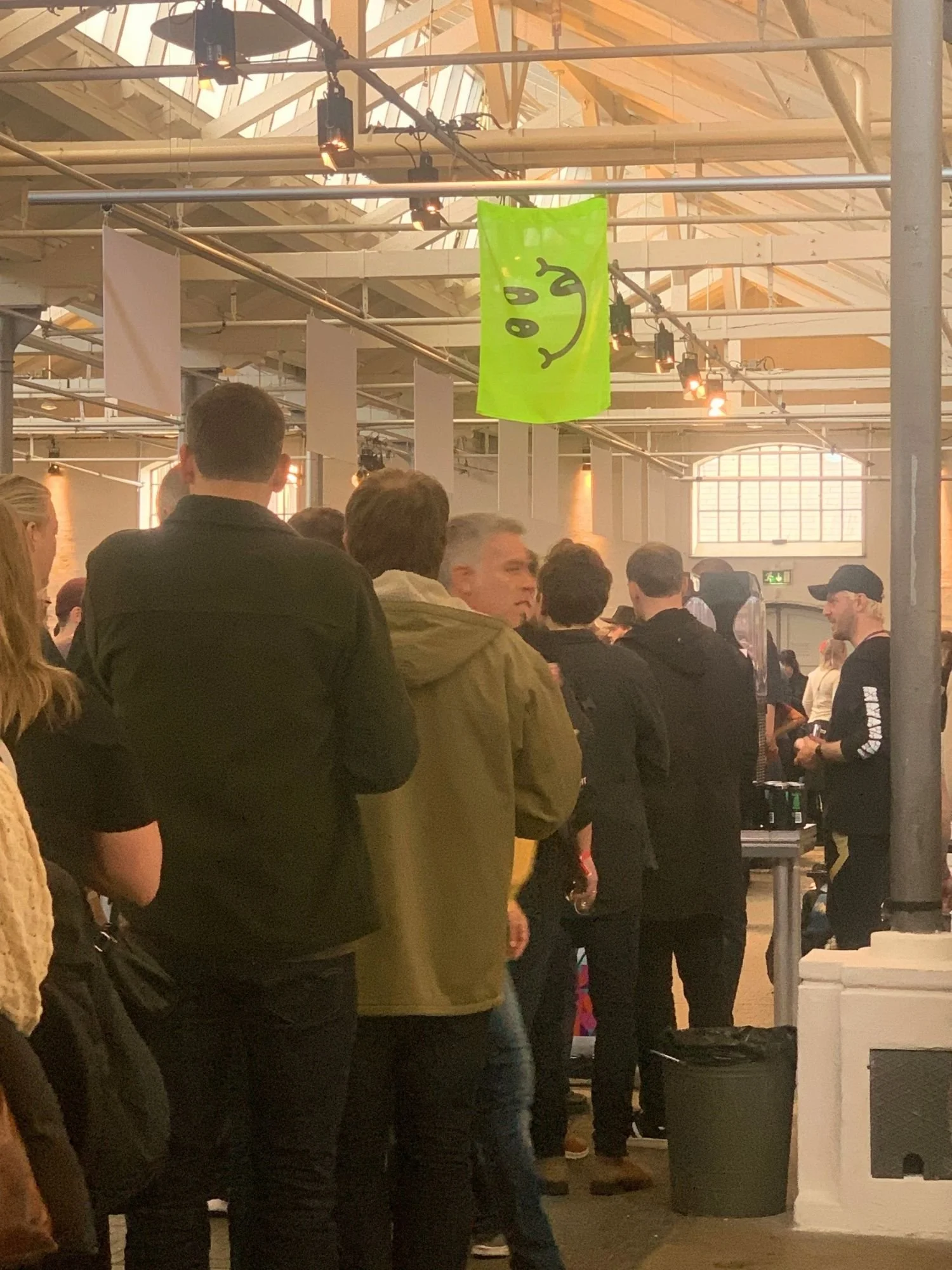 A crowd of people stands in line inside a large venue with high ceilings, exposed beams, and overhead lighting.