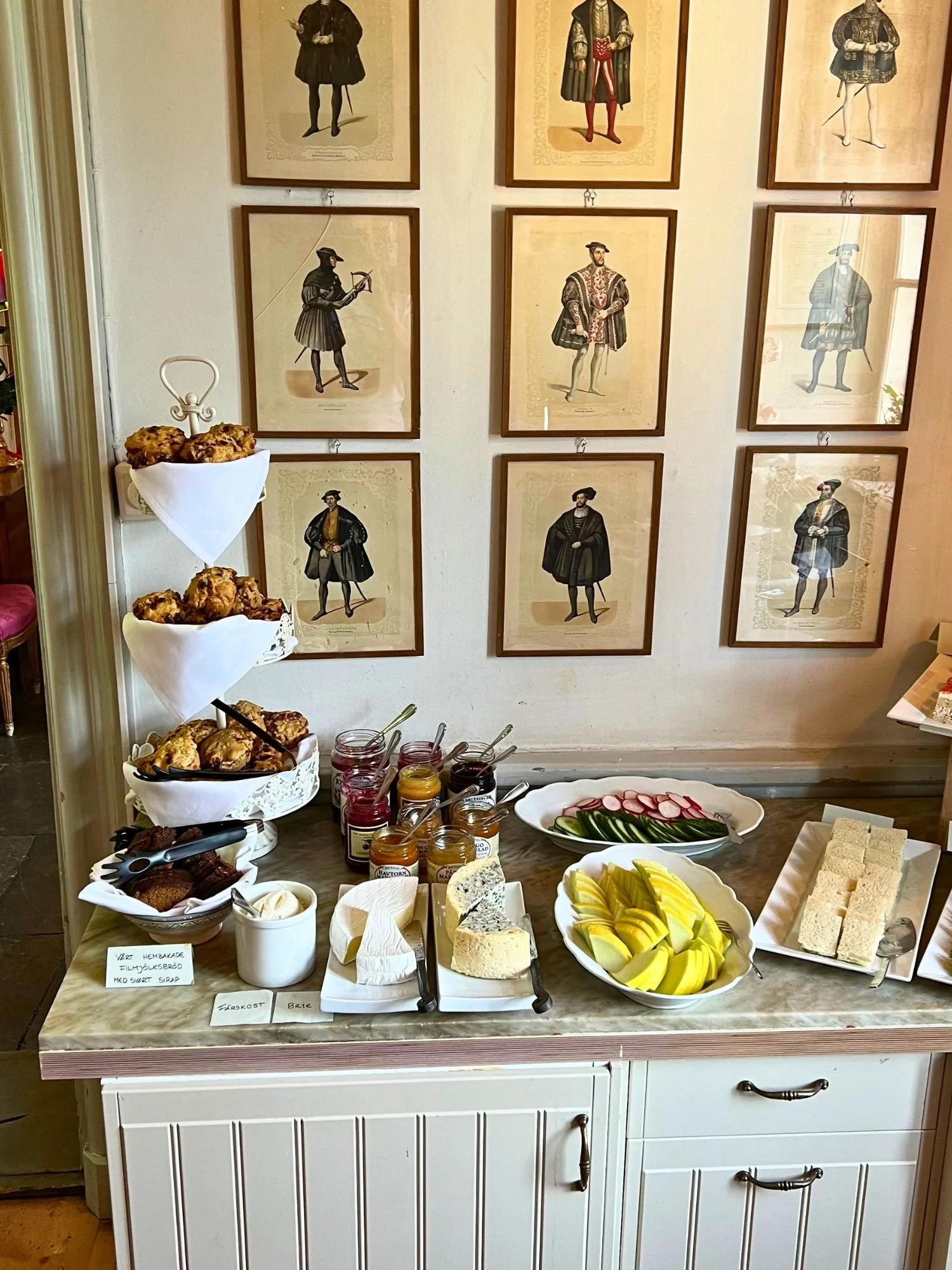 A buffet-style arrangement of savory and sweet items for tea service at Vadstena Klosterhotel. A white three-tiered stand on the left is lined with white napkins and holds a large quantity of golden-brown scones.