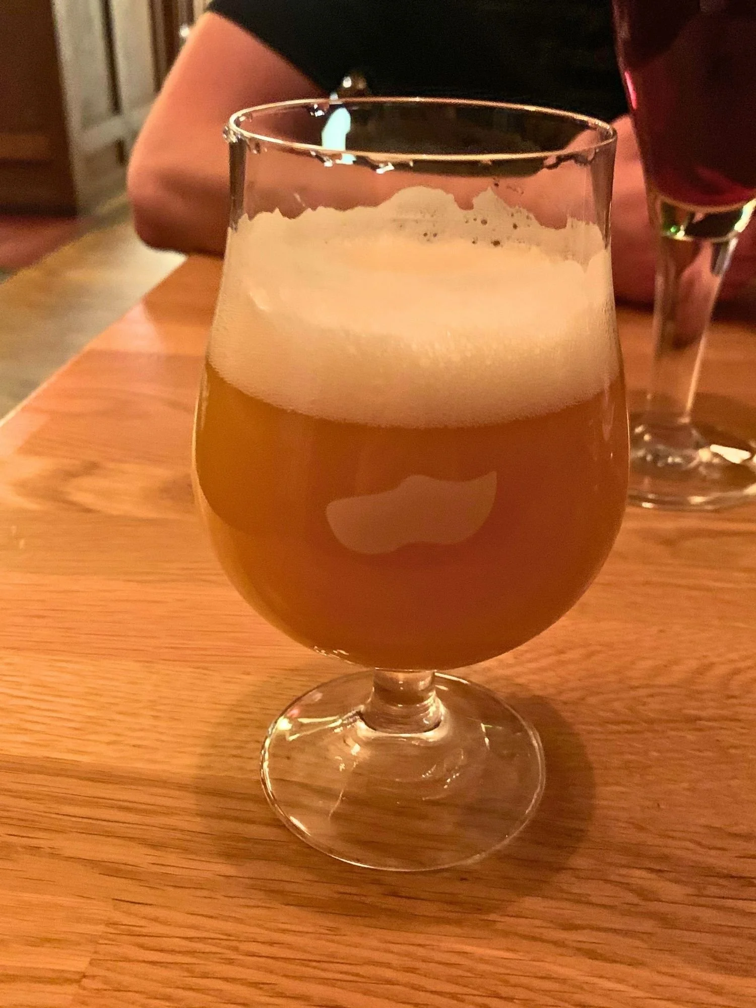 A hazy golden-orange beer with a thick white foam head in a stemmed glass on a wooden table, with another drink and a person in the background.