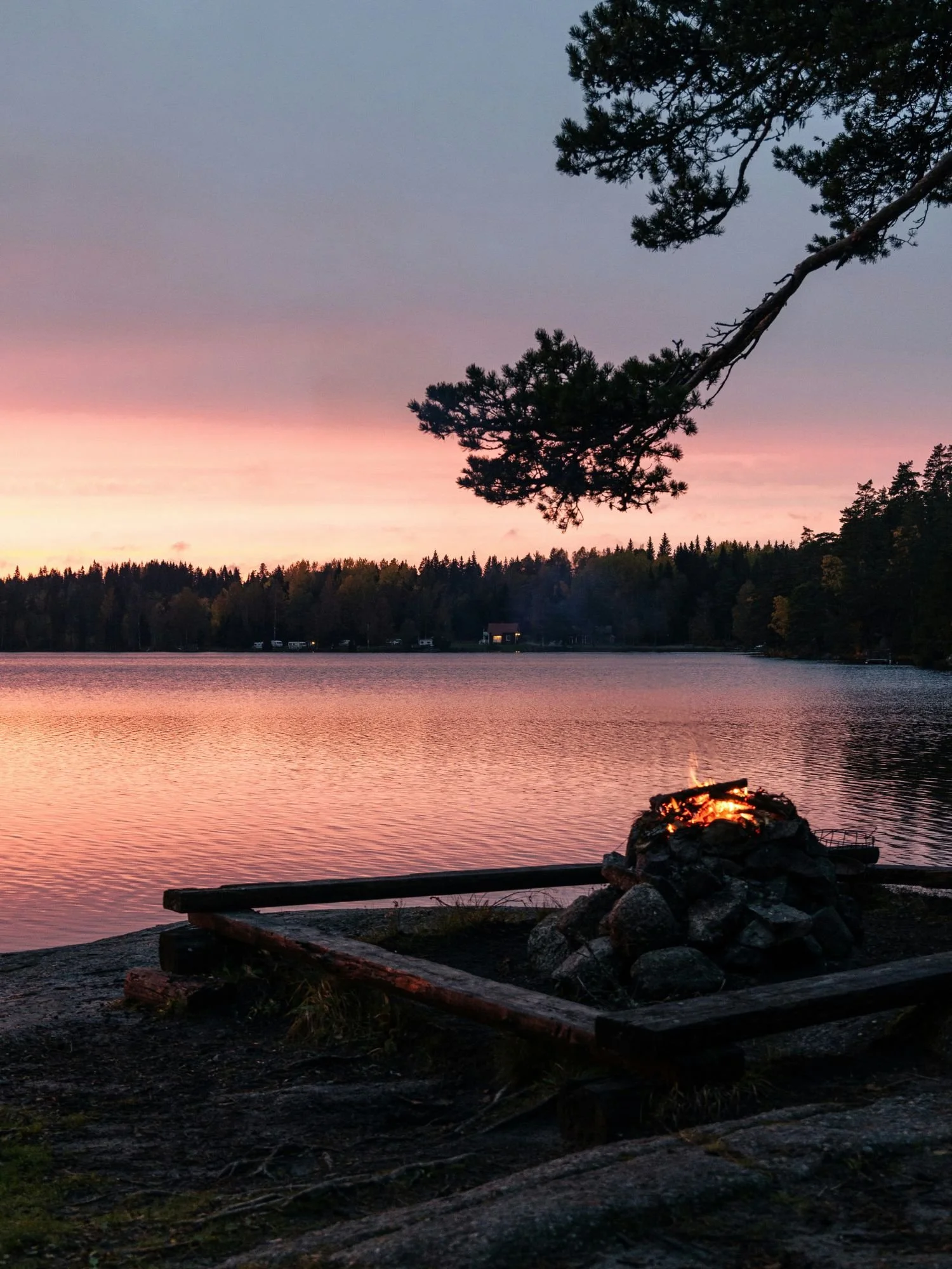 A crackling campfire burns on a stone fire pit built atop a rocky shoreline overlooking a calm lake during a vibrant pink and orange sunset. Simple wooden benches form a square enclosure around the fire pit.