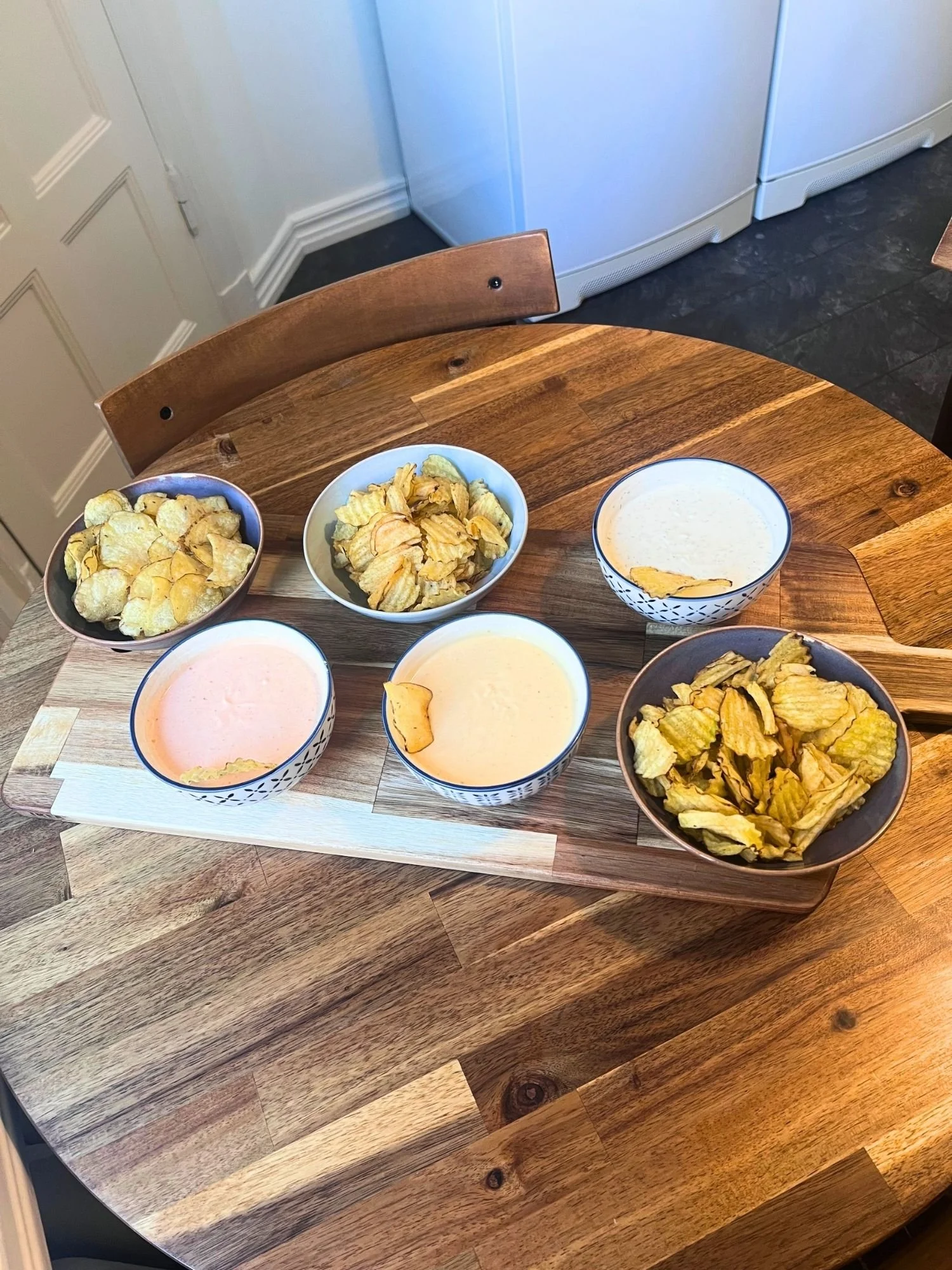 High-angle shot of a wooden serving board set on a round wooden table, holding three bowls of potato chips and three patterned ceramic bowls of creamy dips.