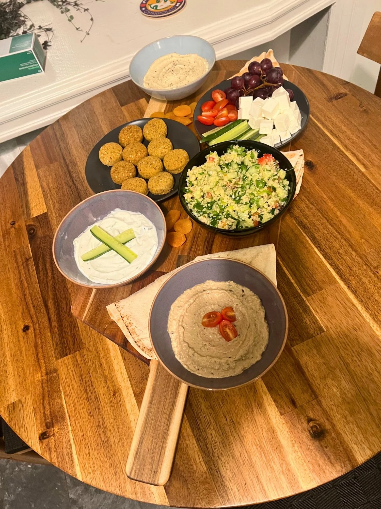 High-angle shot of a wooden board on a round wooden table, set with a Mediterranean mezze spread including falafel balls, tabbouleh, hummus, tzatziki, cubes of feta cheese, cherry tomatoes, cucumber slices, red grapes, dried apricots, and pita bread.