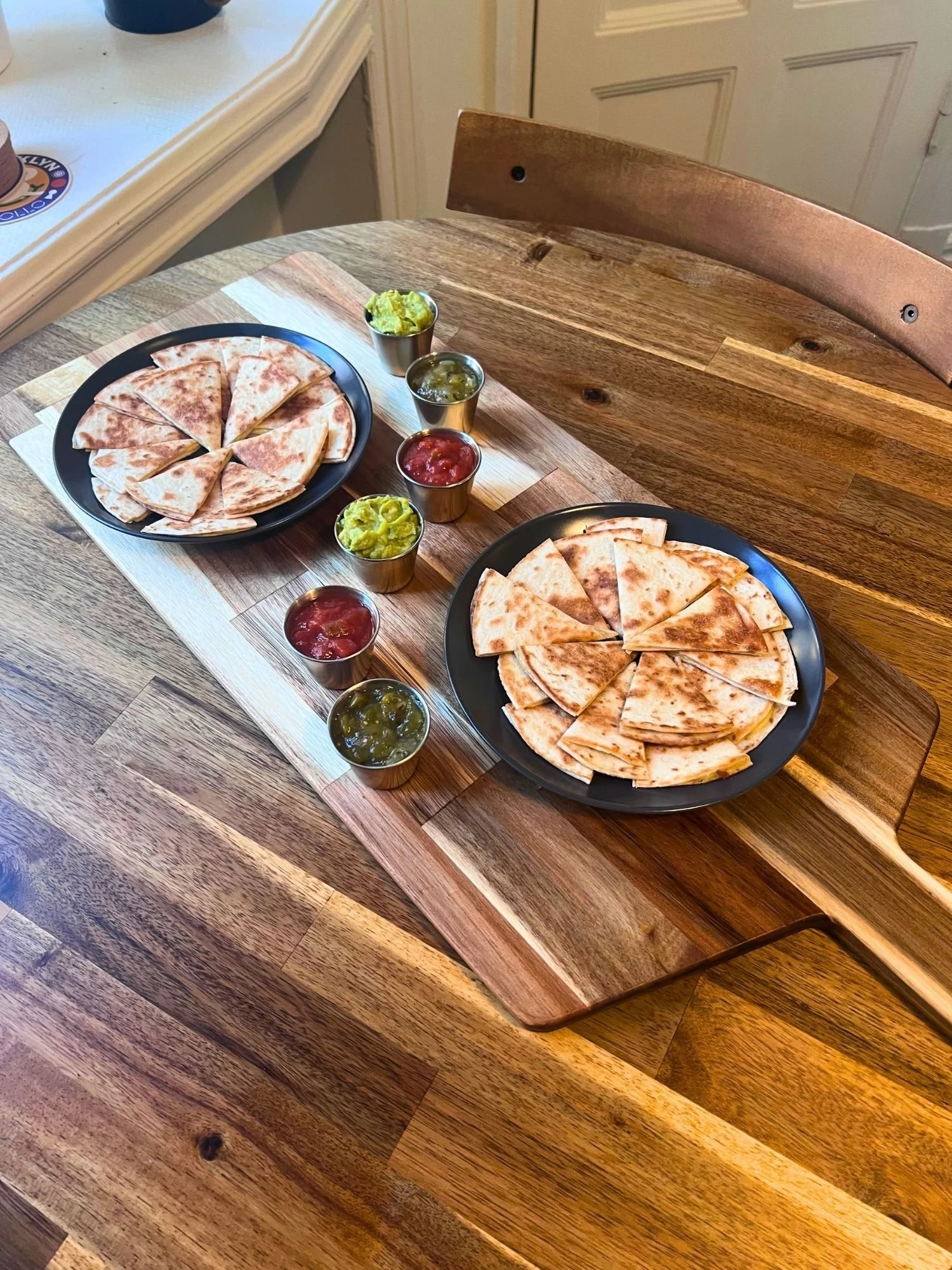 High-angle shot of a long wooden serving board on a round wooden table, set with two dark plates of sliced quesadilla wedges and small metal ramekins filled with guacamole and salsa.