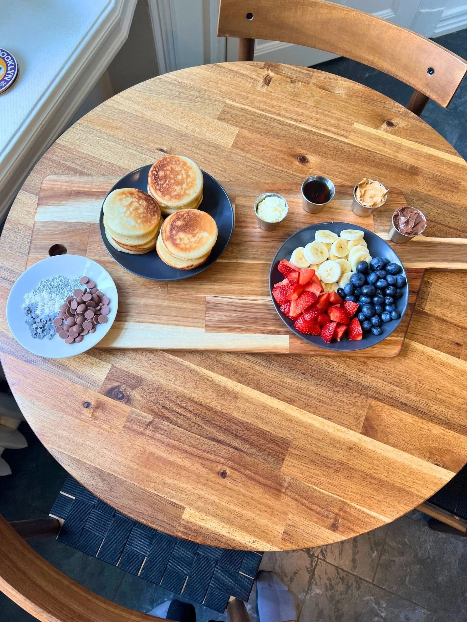 Overhead view of a wooden serving board on a round wooden table, set with plates of mini pancakes, fresh sliced strawberries, bananas, blueberries, chocolate chips, sprinkles, and small cups of butter, syrup, peanut butter, and chocolate spread.