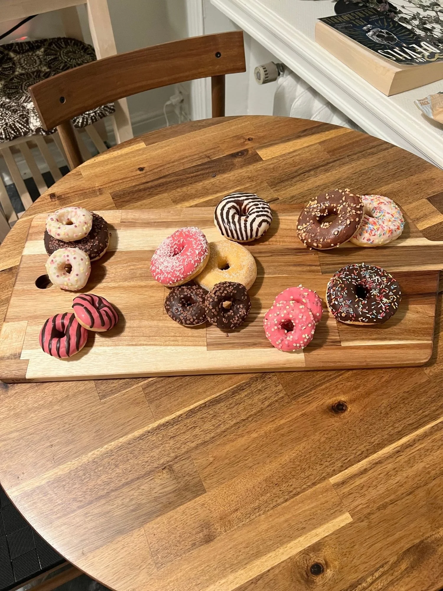 A wooden serving board on a round wooden table, arranged with an assortment of full-size and mini decorated donuts.