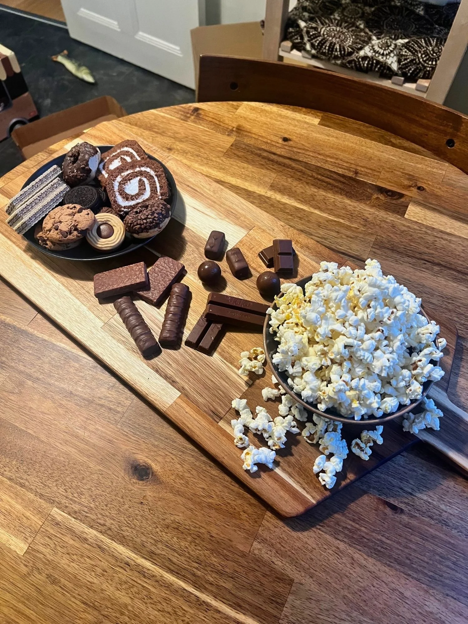 A wooden table set with a serving paddle holding a dark gray plate of assorted cookies, a bowl of popcorn with pieces of popcorn scattered around it, and various chocolate bars and candies arranged in the center.