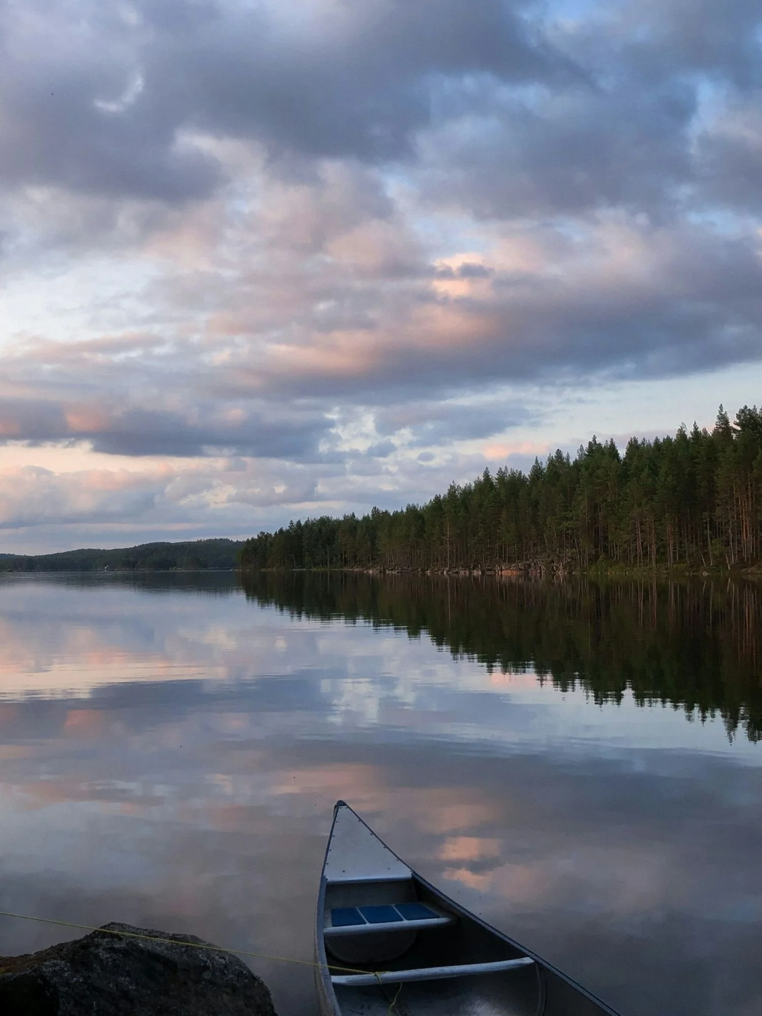 A scenic, wide shot from the perspective of a shoreline looking out over a perfectly calm, glassy lake at dawn. In the foreground, the bow of an aluminum canoe is visible, resting by a dark rock.