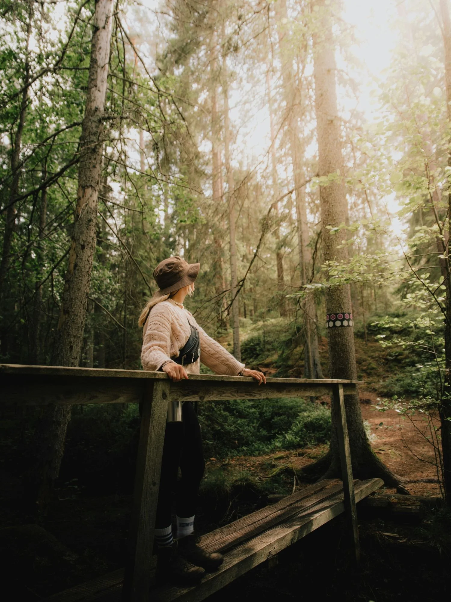 A woman wearing a brown bucket hat, a chunky beige knit sweater, dark trousers, and hiking boots stands on a rustic wooden footbridge in Tiveden National Park.