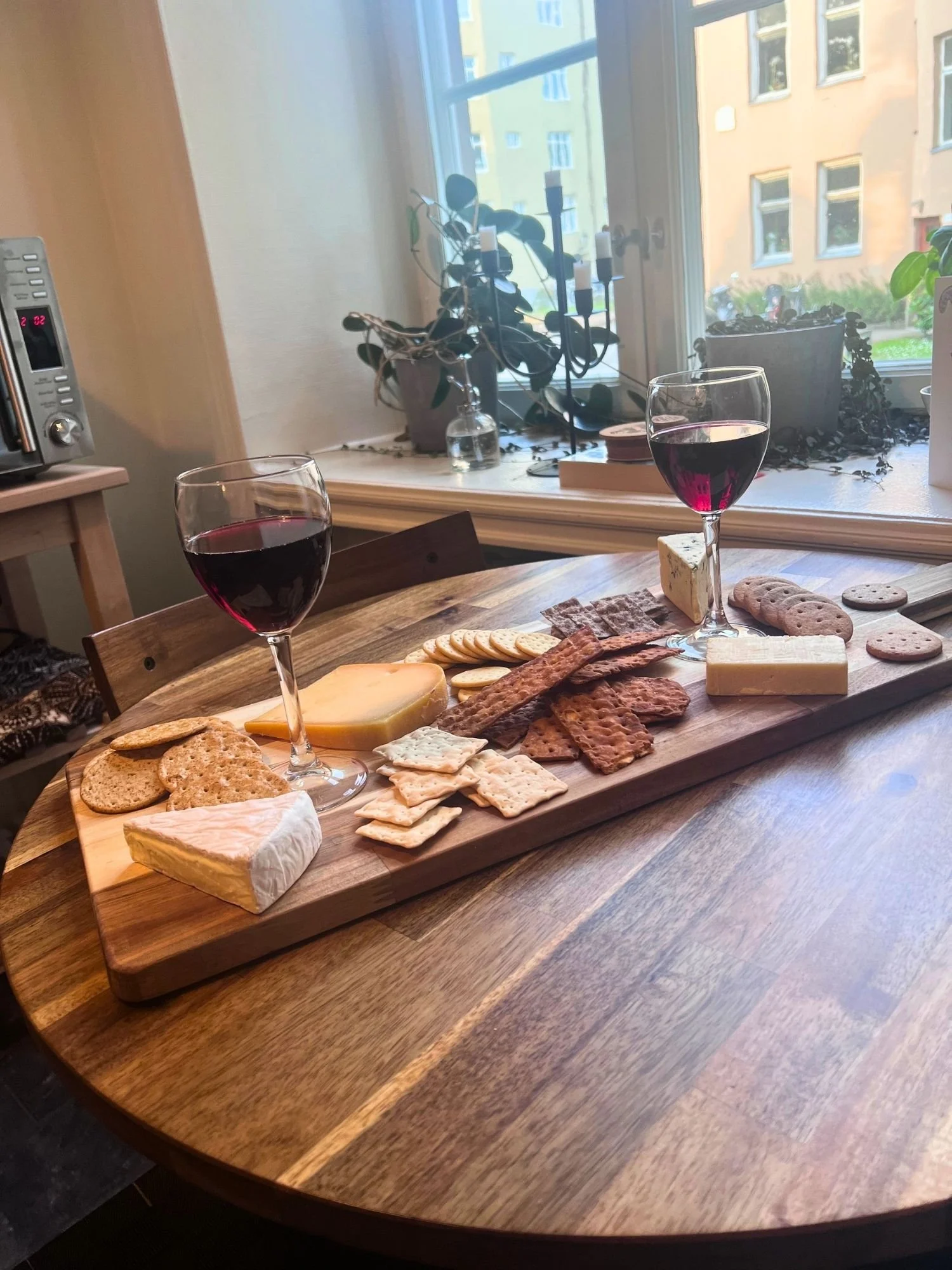 A wooden charcuterie board filled with assorted cheeses and crackers set on a round wooden table, flanked by two glasses of red wine near a window.