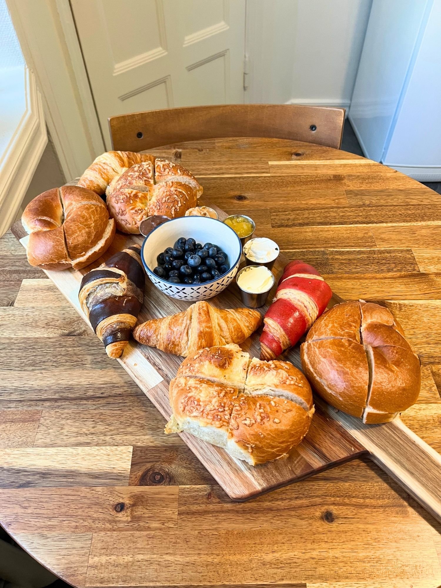 High-angle shot of a wooden serving board on a round wooden table, arranged with a variety of croissants and bread rolls, a small bowl of fresh blueberries, and tiny metal ramekins containing spreads and butter.