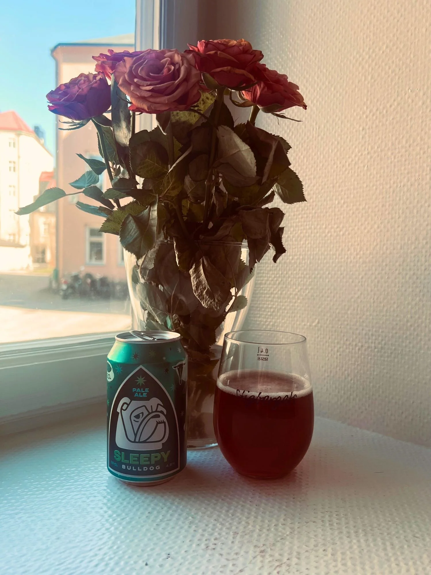 A can and glass of pale ale on a windowsill beside a vase of pale orange roses, with warm sunlight illuminating the scene and buildings visible outside the window.