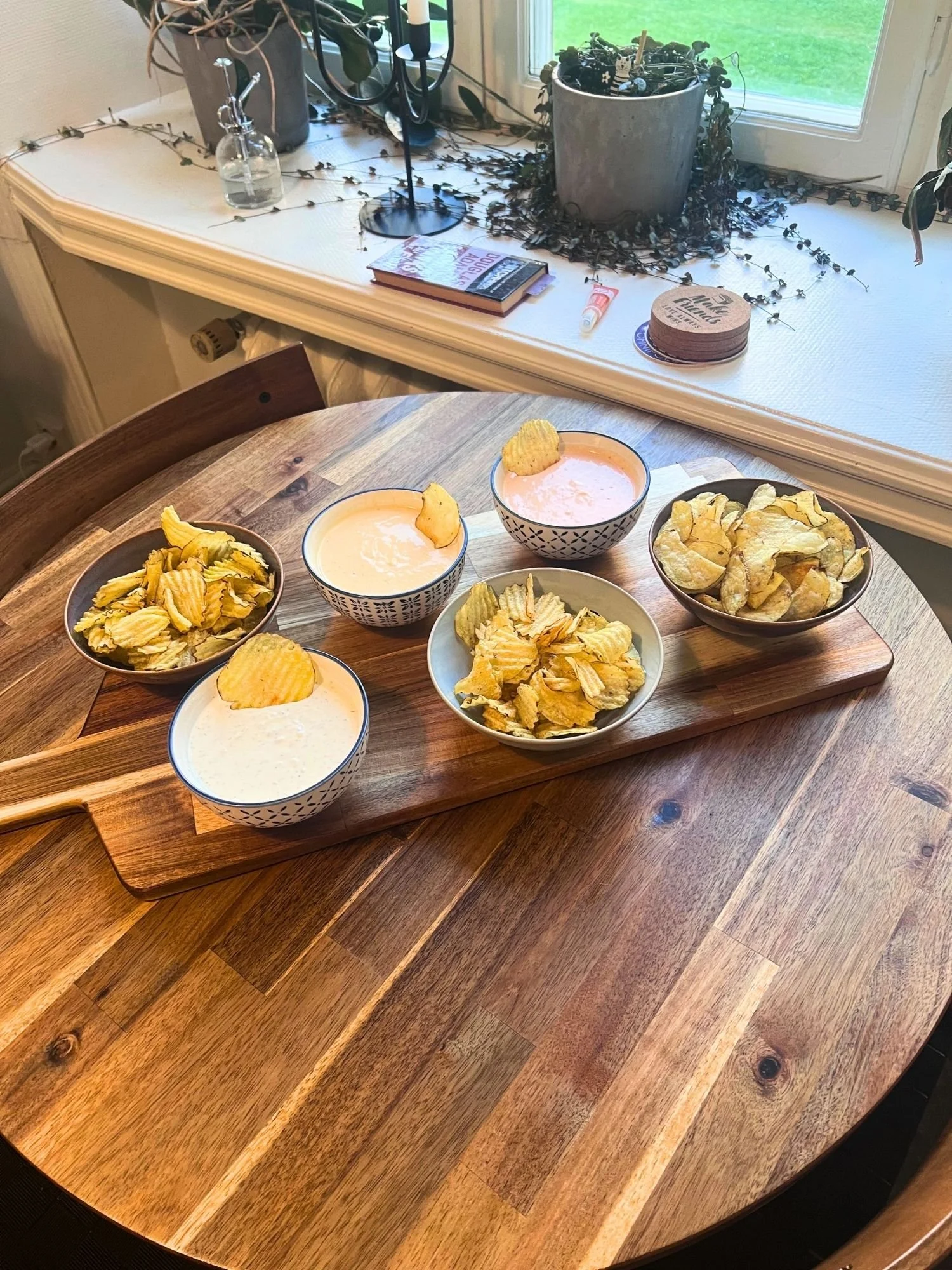 A wooden table set with a serving paddle holding three bowls of potato chips and three patterned bowls of creamy dipping sauces, each with a chip resting inside.
