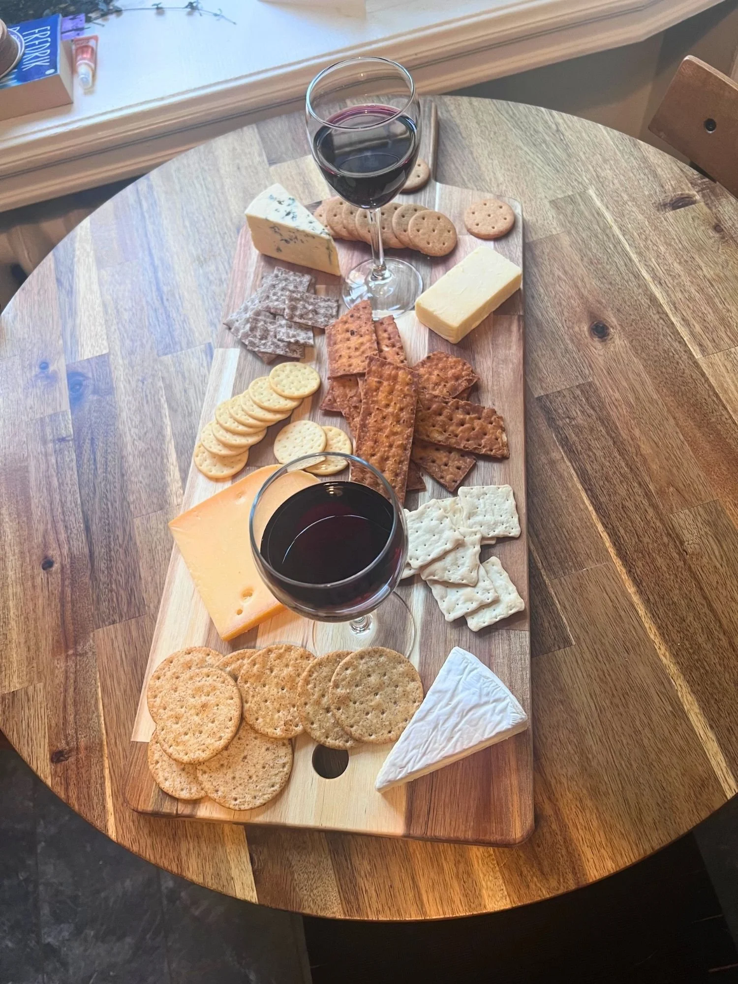 High-angle shot of a large wooden board on a round table, filled with an assortment of cheeses, various crackers, and two glasses of red wine.