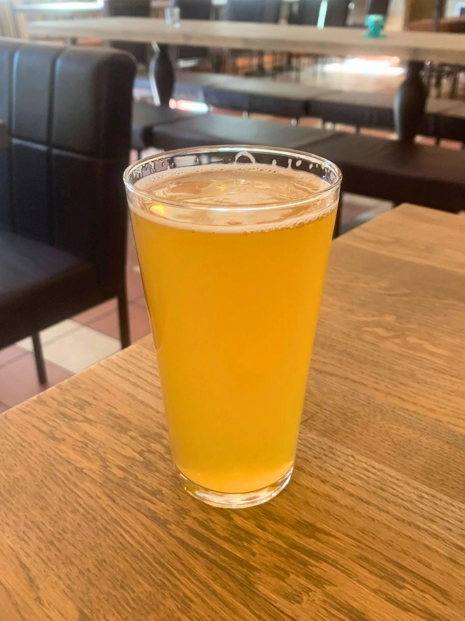 A full pint glass of hazy, golden-orange draft beer sitting on a light wooden table. A black leather chair and empty restaurant tables with bench seating are visible in the blurred background.