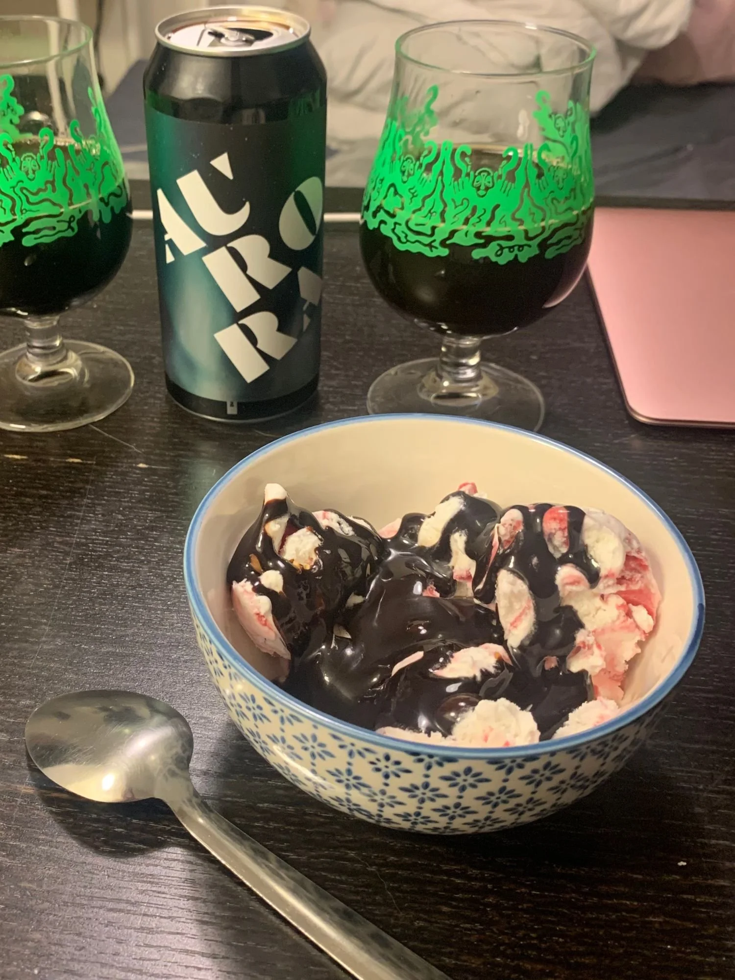 A bowl of ice cream with chocolate sauce on a dark table next to a can of Spike Brewery Aurora beer and two stemmed glasses filled with dark stout.