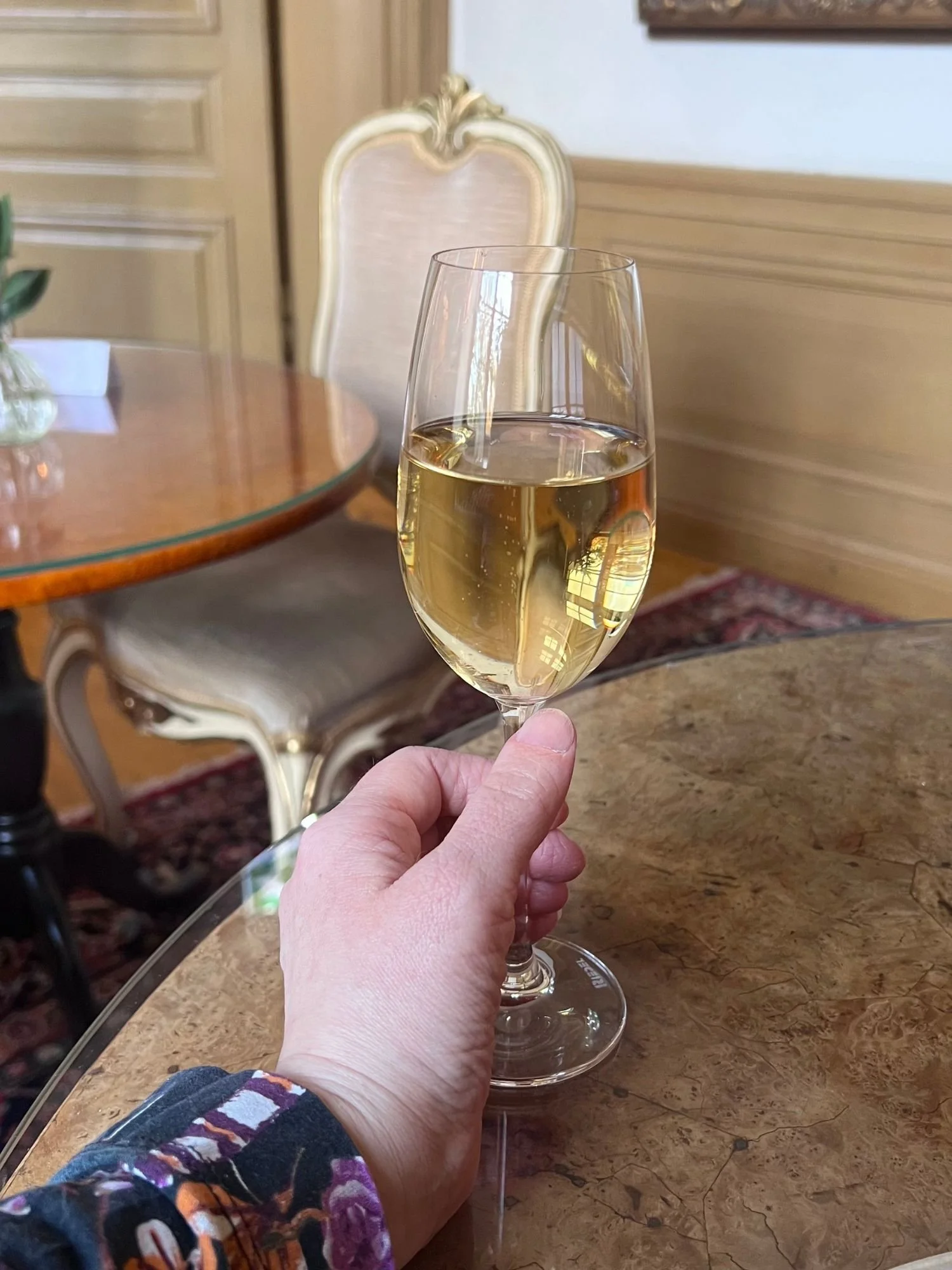 A hand holding a glass of champagne at afternoon tea at Vadstena Klosterhotel. The glass is held over a marble-topped table with a polished, brown-and-tan patterned finish.