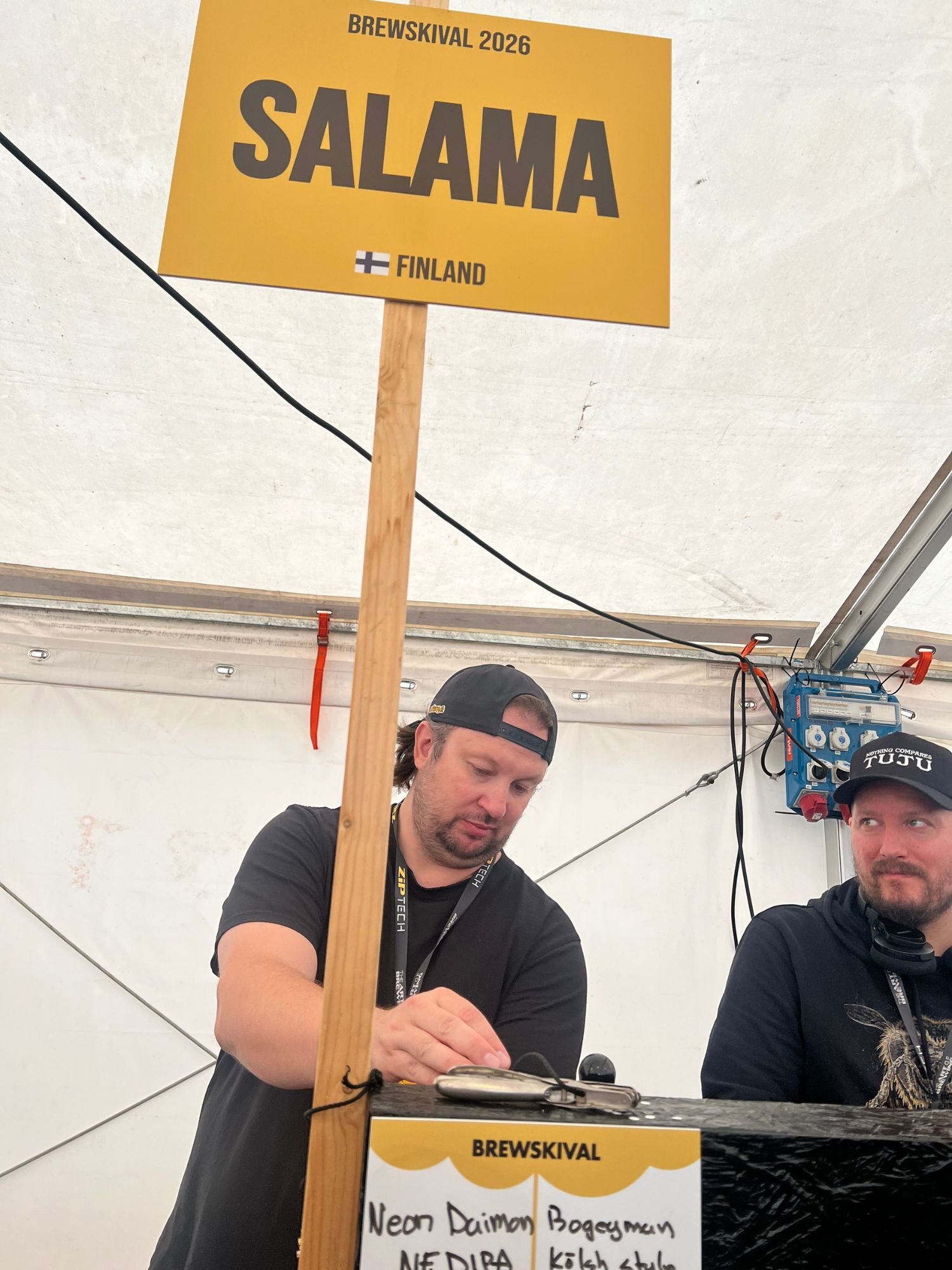 Two men working behind a festival counter beneath a yellow sign with text "Salaam - Finland." The man on the left wears a black shirt and backward cap while adjusting equipment on the counter, and the man on the right wears a black hoodie and a cap.