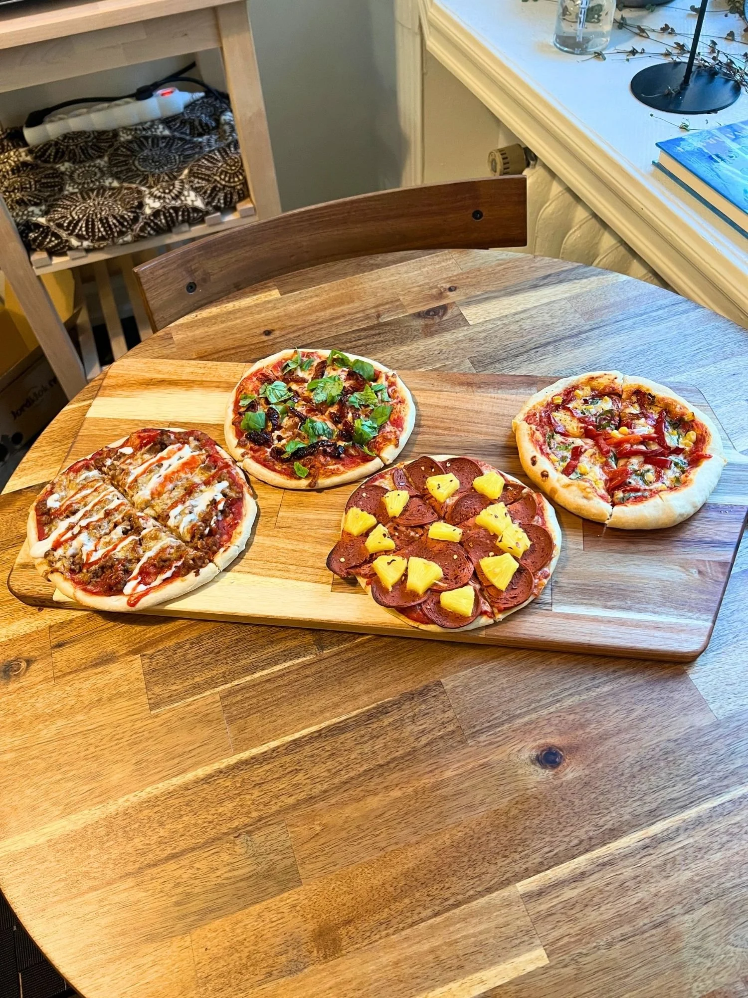 A wooden serving board on a round wooden table, displaying four different personal-sized pizzas with various toppings.