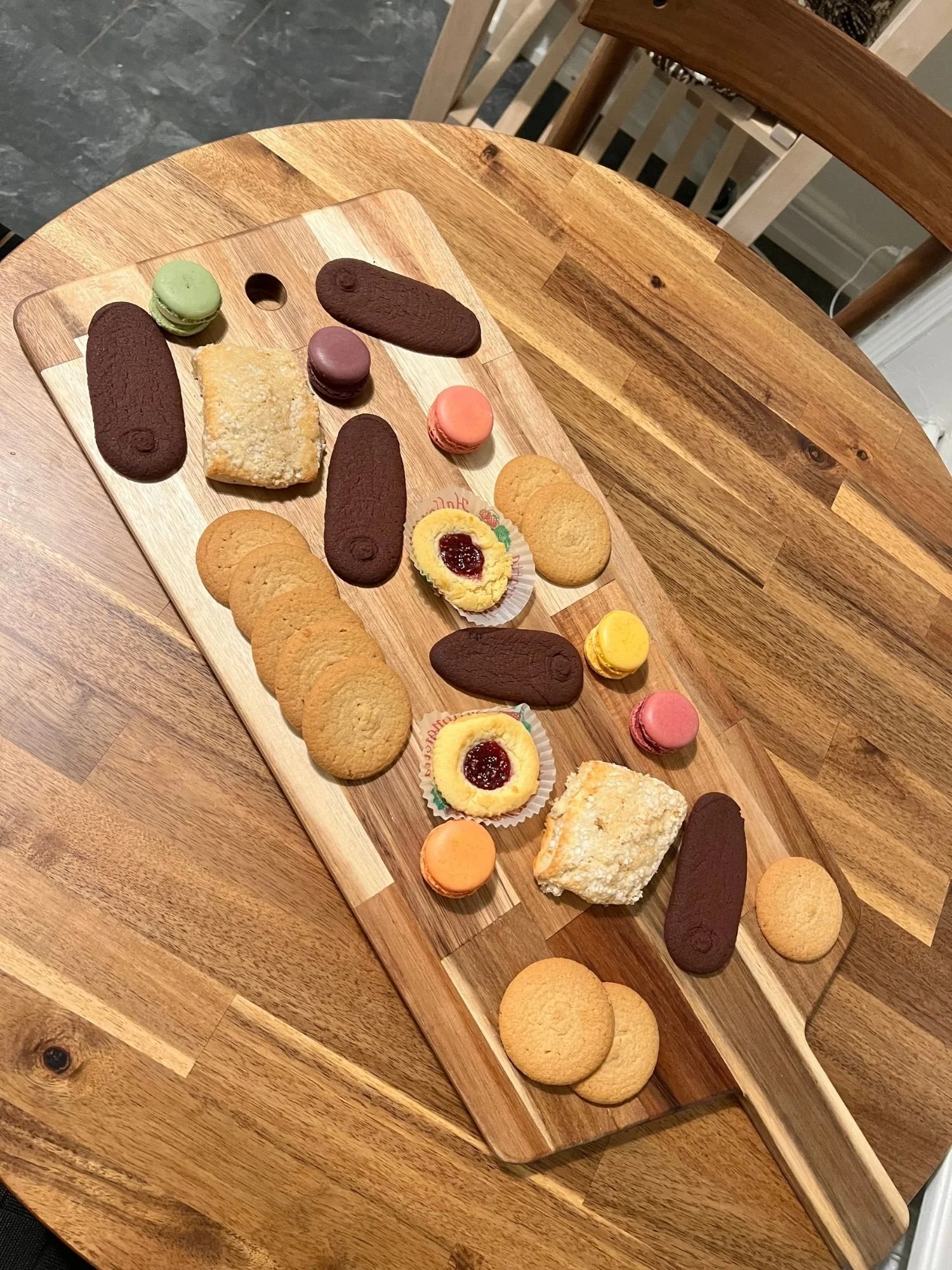 A long wooden cutting board rests on a round wooden table, arranged with a variety of cookies, pastries, and colorful macarons.