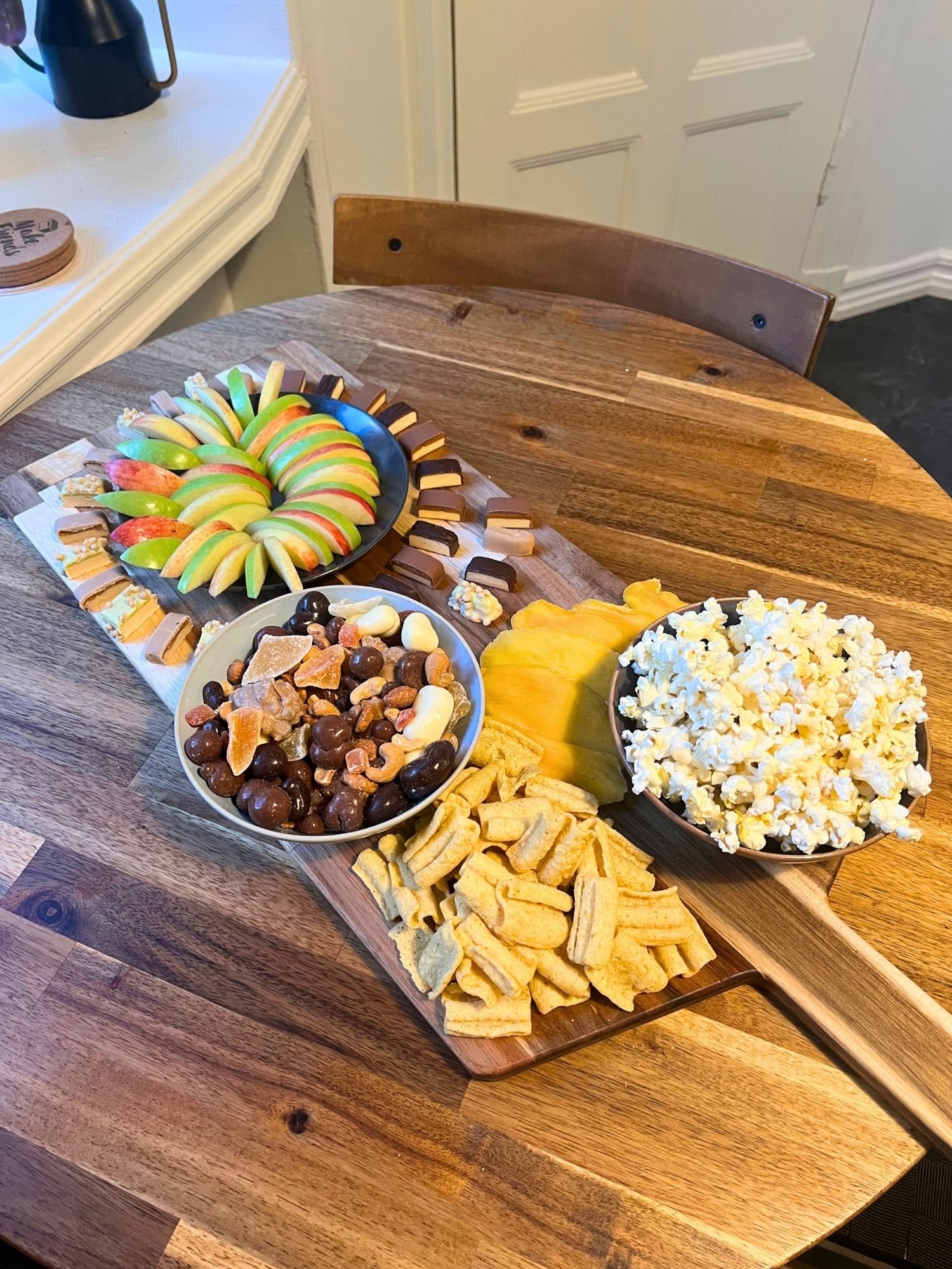 High-angle, diagonal view of a wooden serving board set on a round wooden table, holding bowls of popcorn and trail mix, crunchy snacks, dried mango, and sliced apples surround protein bar bites.