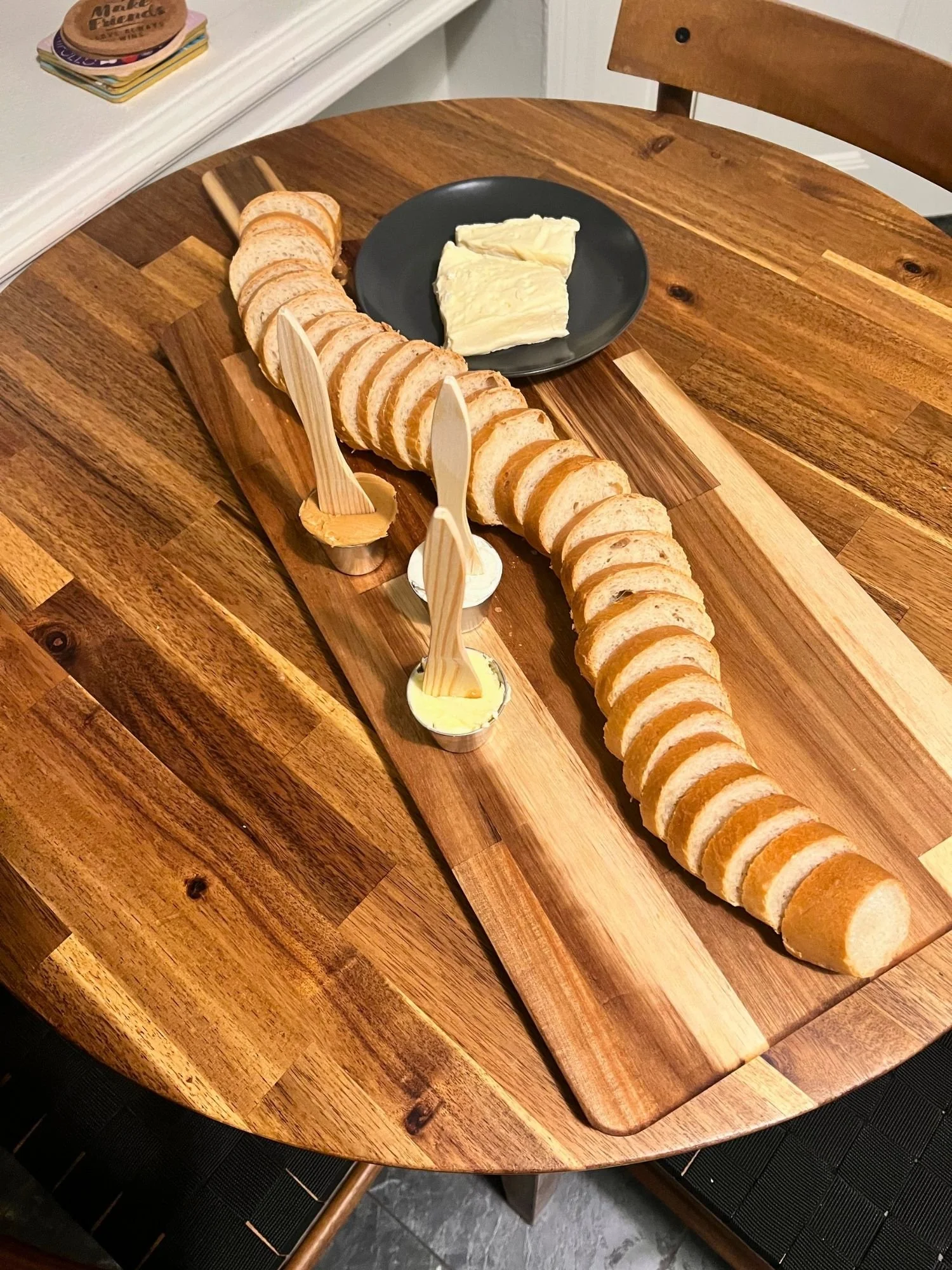 High-angle shot of a long wooden serving board on a round wooden table, featuring a long sliced baguette snaking down the middle, accompanied by small cups of spreads with wooden utensils and a dark plate holding slices of soft cheese.