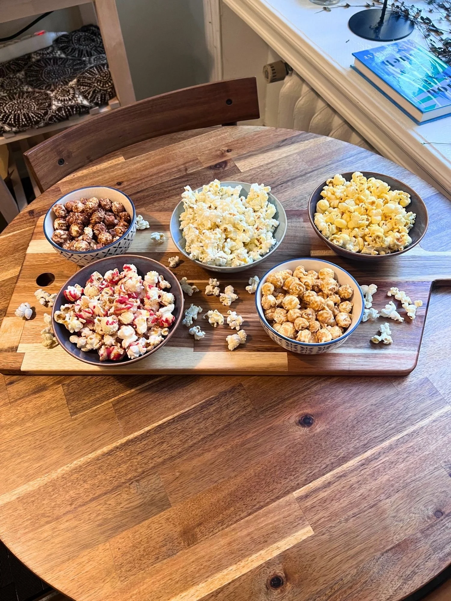 High-angle view of a wooden serving board set on a round wooden table, holding five ceramic bowls with different varieties of gourmet popcorn.