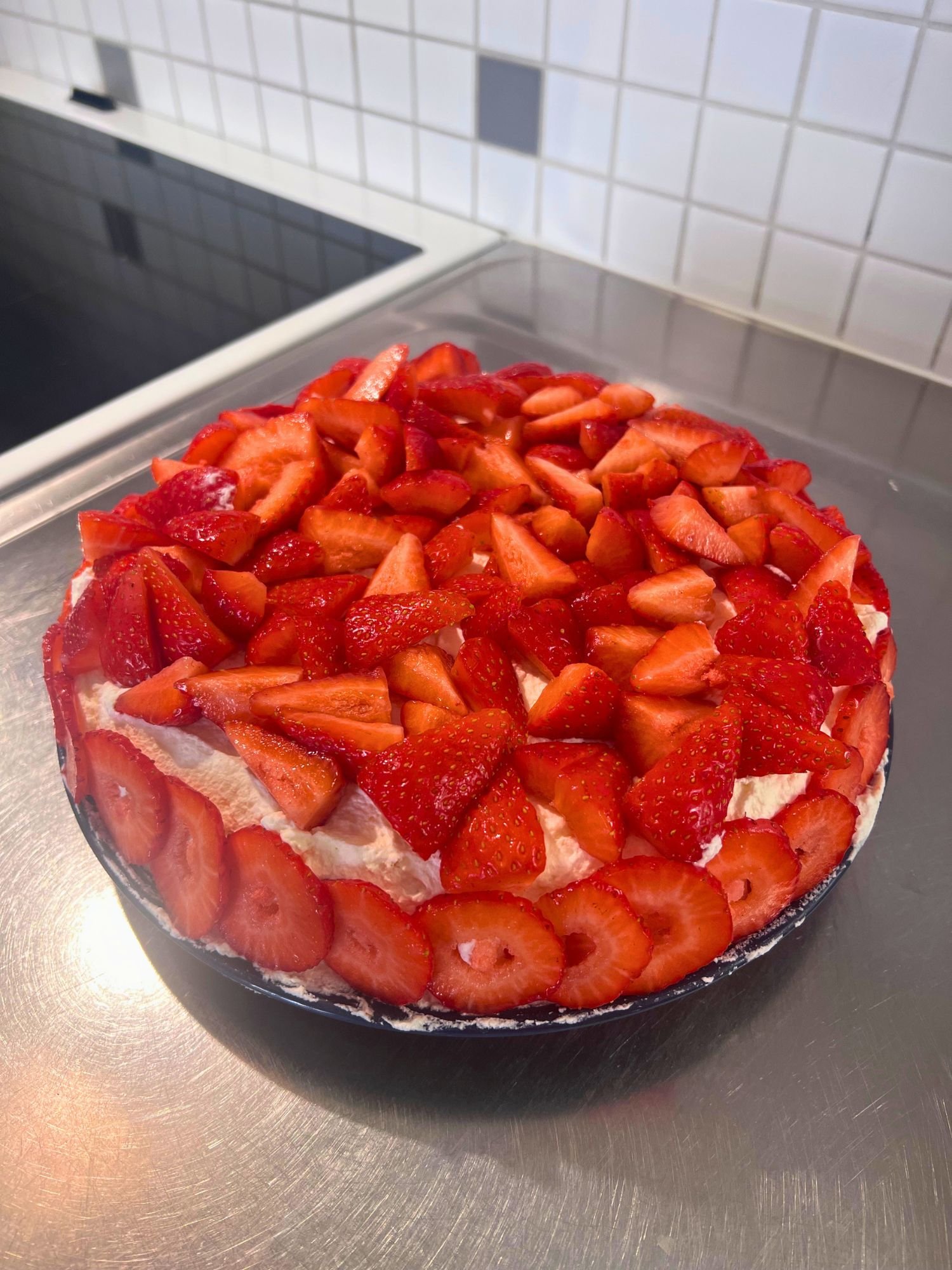 Homemade Swedish summer cake topped with whipped cream and fresh strawberries, arranged on a round cake base with sliced strawberries decorating the sides. The cake sits on a stainless steel countertop in a bright kitchen.