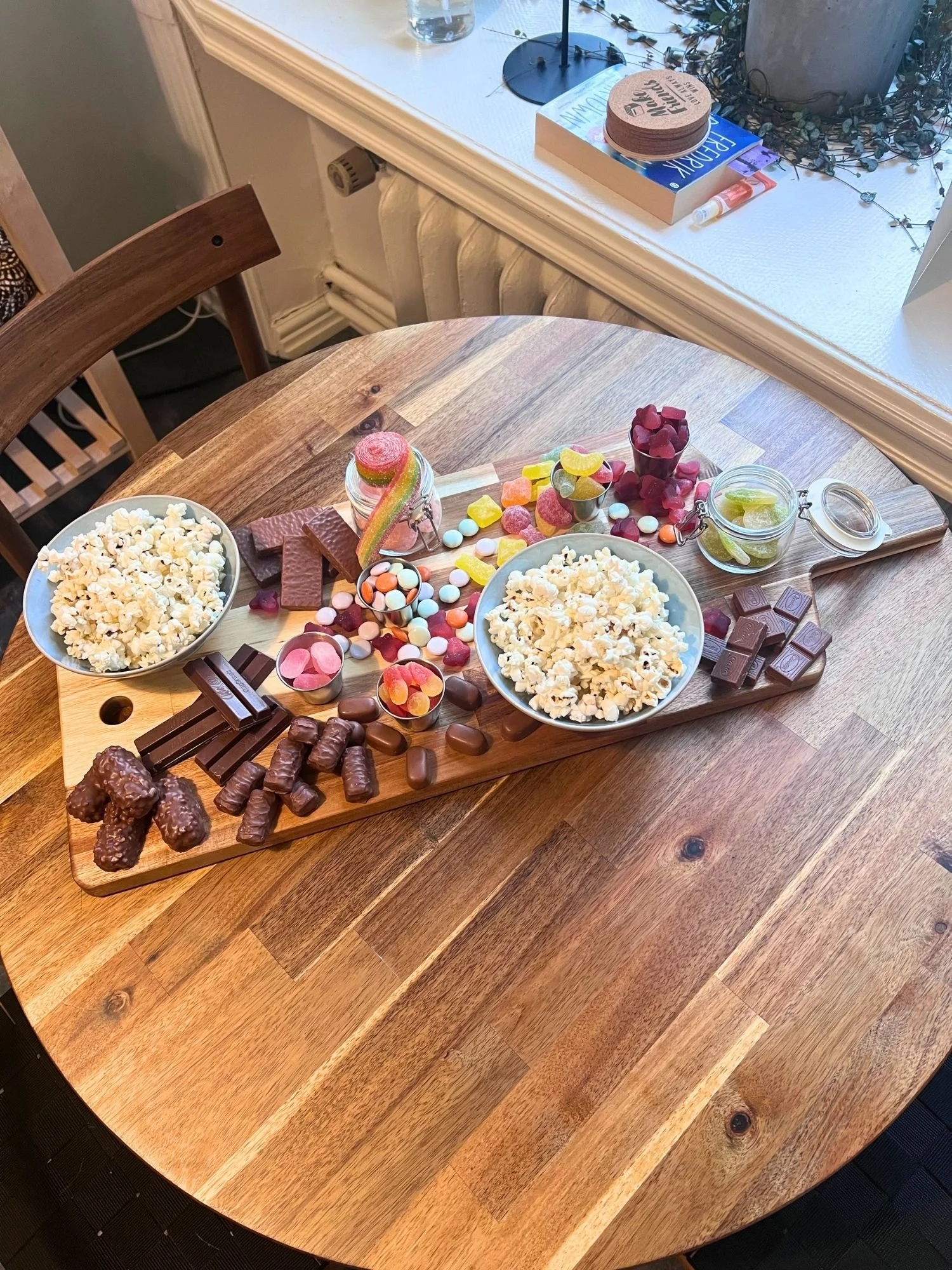 A wooden table set with a large snack board loaded with two bowls of popcorn, glass jars of colorful gummy candies, chocolate bars, and candy-coated chocolates.