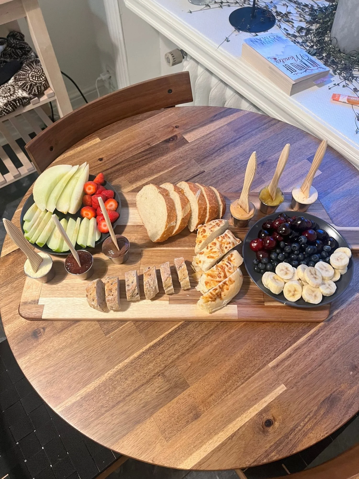 A large rectangular wooden charcuterie board sits atop a round wooden table, laden with an array of sliced breads, fresh fruits, and small cups of spreads.