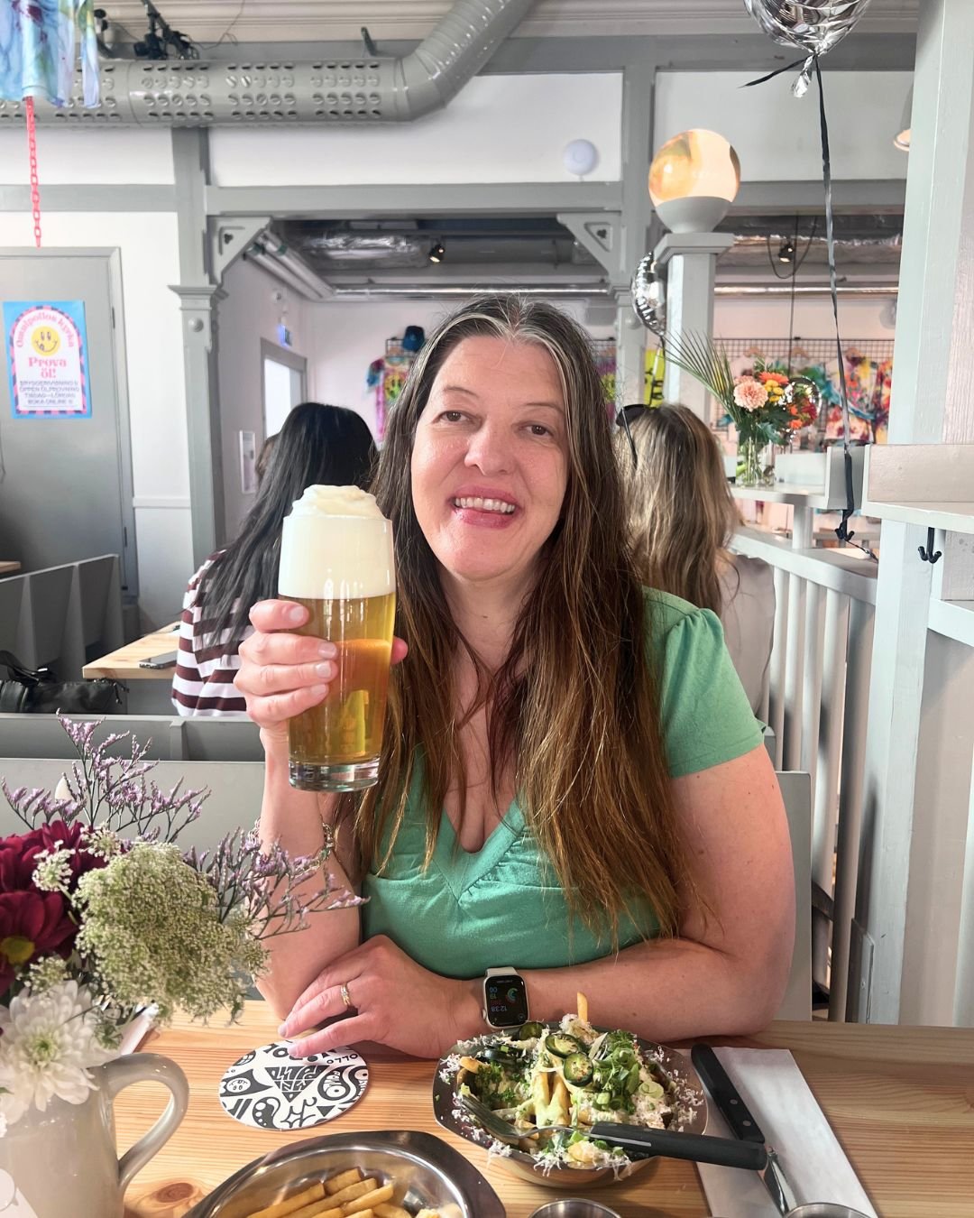 A woman seated at a brewery restaurant holding a glass of pale craft beer with a thick foamy head. There are fries and flowers on the wooden table, with the bright, modern brewery interior visible in the background.