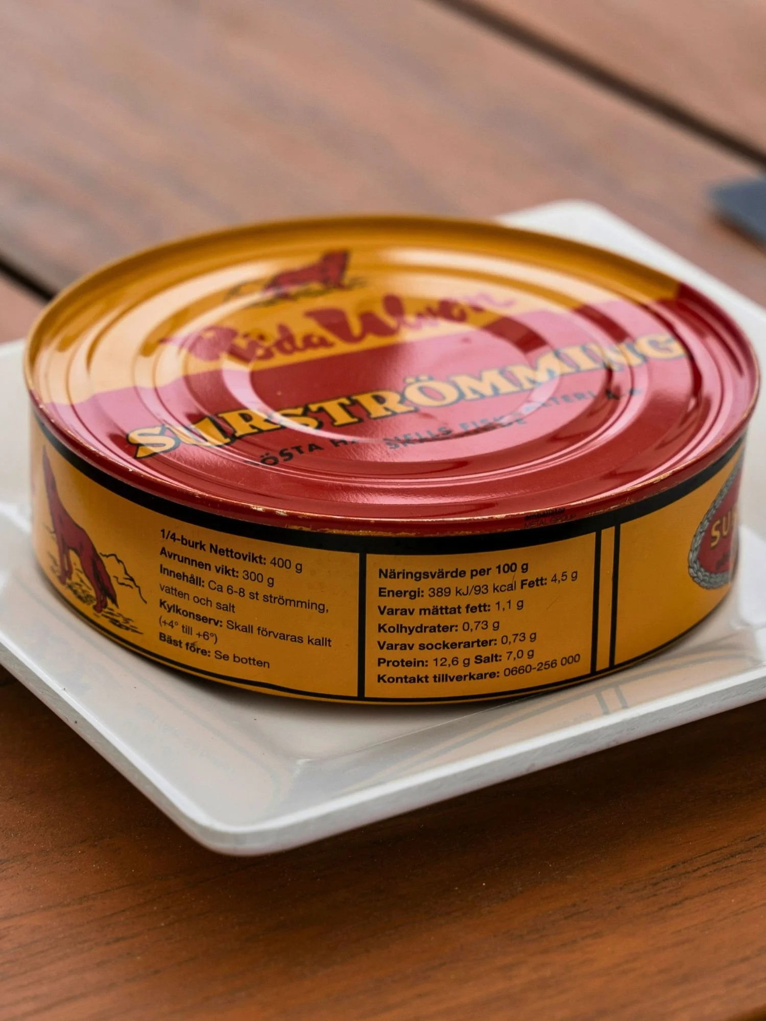 A sealed tin of Röda Ulven Surströmming (fermented Baltic Sea herring) on a small white square plate. The cylindrical tin features a classic yellow and red design with a red wolf logo.