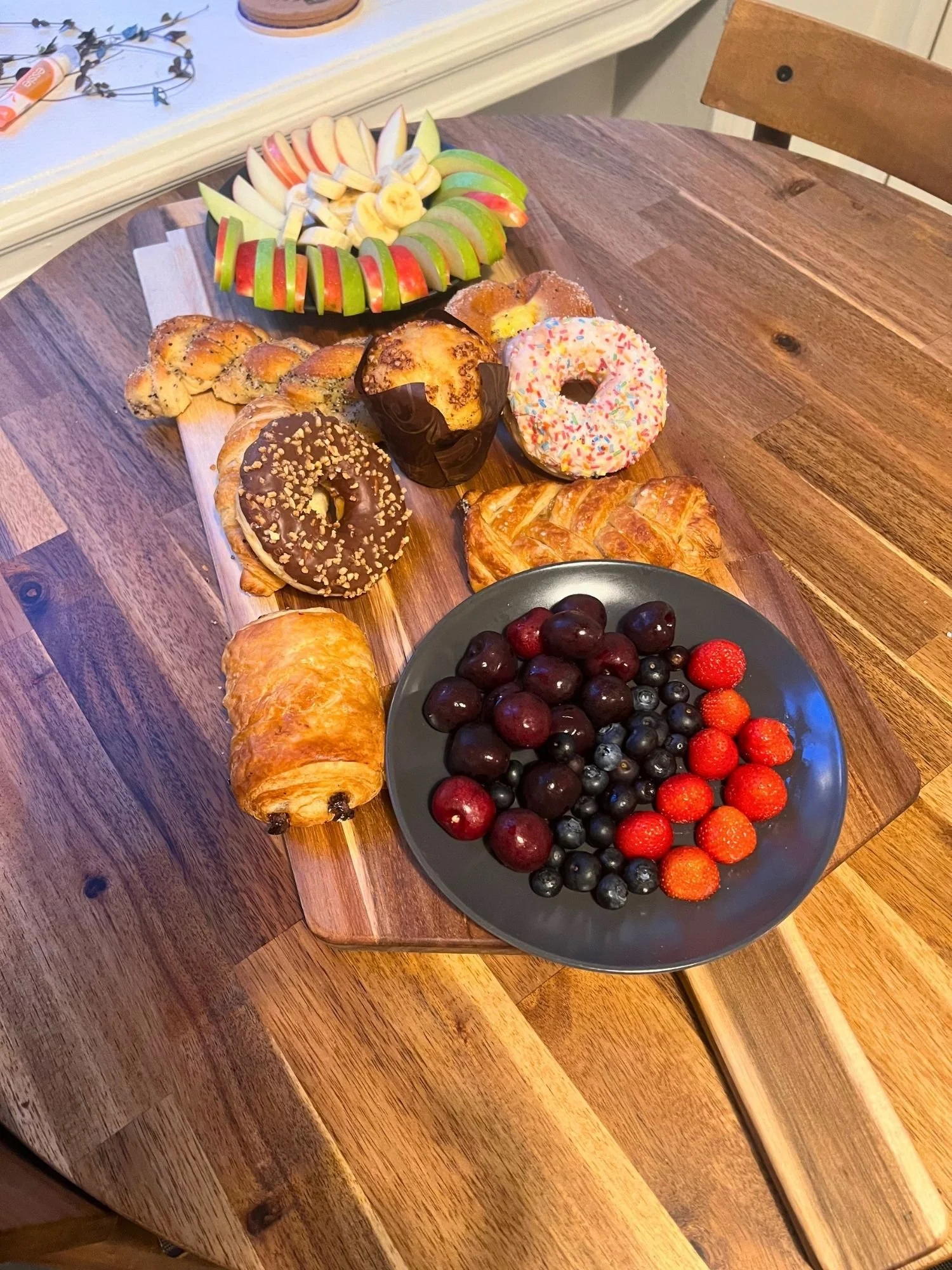 A long wooden serving board on a wooden table, arranged with sweet pastries and fresh fruit.